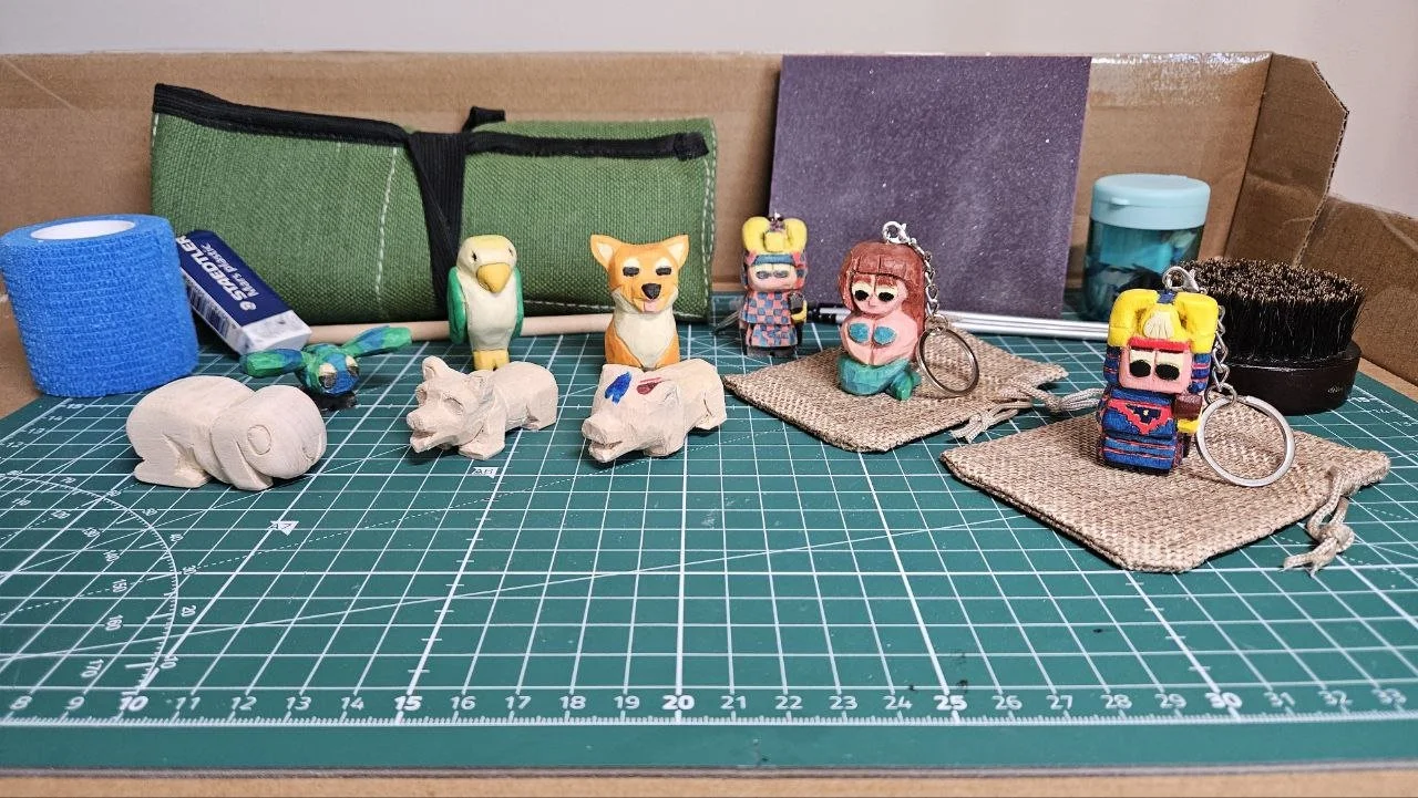 Una collezione di gadget e oggetti artigianali su una superficie di taglio verde, tra cui figurine, chiavette, rotoli di nastro adesivo, un pennello, un rotolo di cartoncino e blocchi di schiuma, con una scatola di cartone come sfondo.