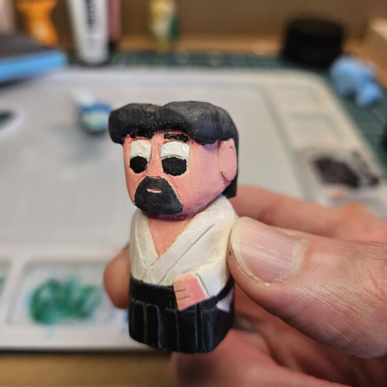 Statua di legno dipinta di un personaggio con capelli e barba neri, pelle rosa, vestito bianco e cintura nera, in uno stile cartoon.