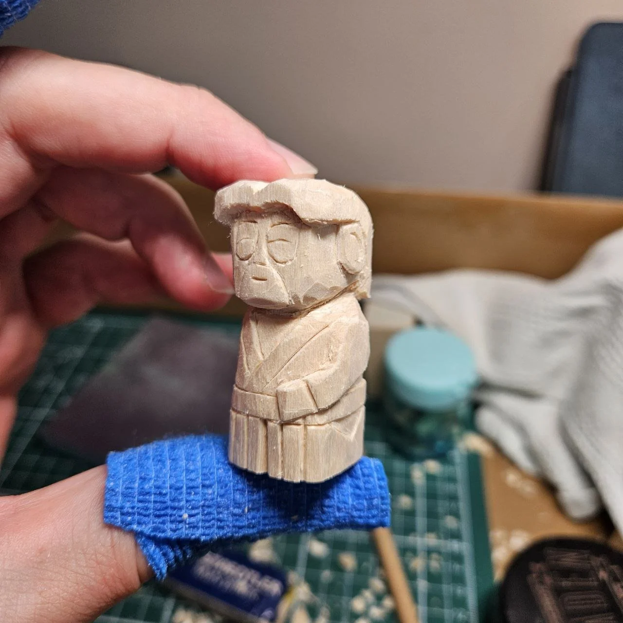 Scultura in legno di un personaggio stilizzato con capelli, occhi e abbigliamento semplice, tenuto con una mano.