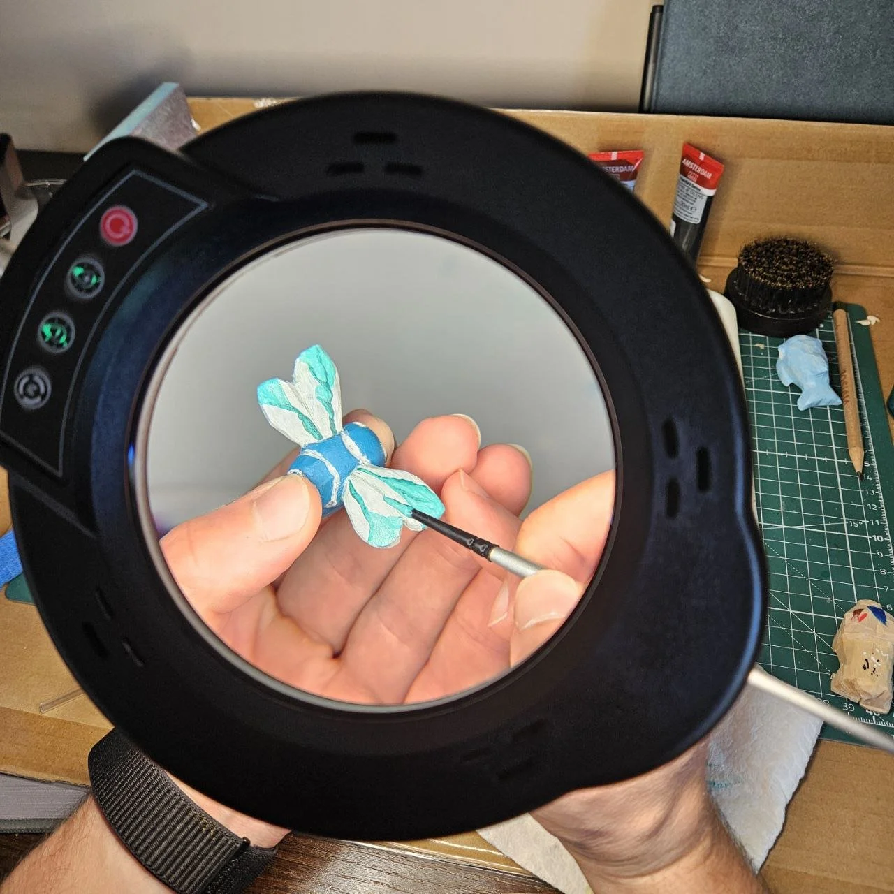 Una mano dipinge dettagli di una farfalla di carta con un piccolo pennello, vista attraverso un'icona di un visore di realtà virtuale in un ambiente di lavoro artistico.