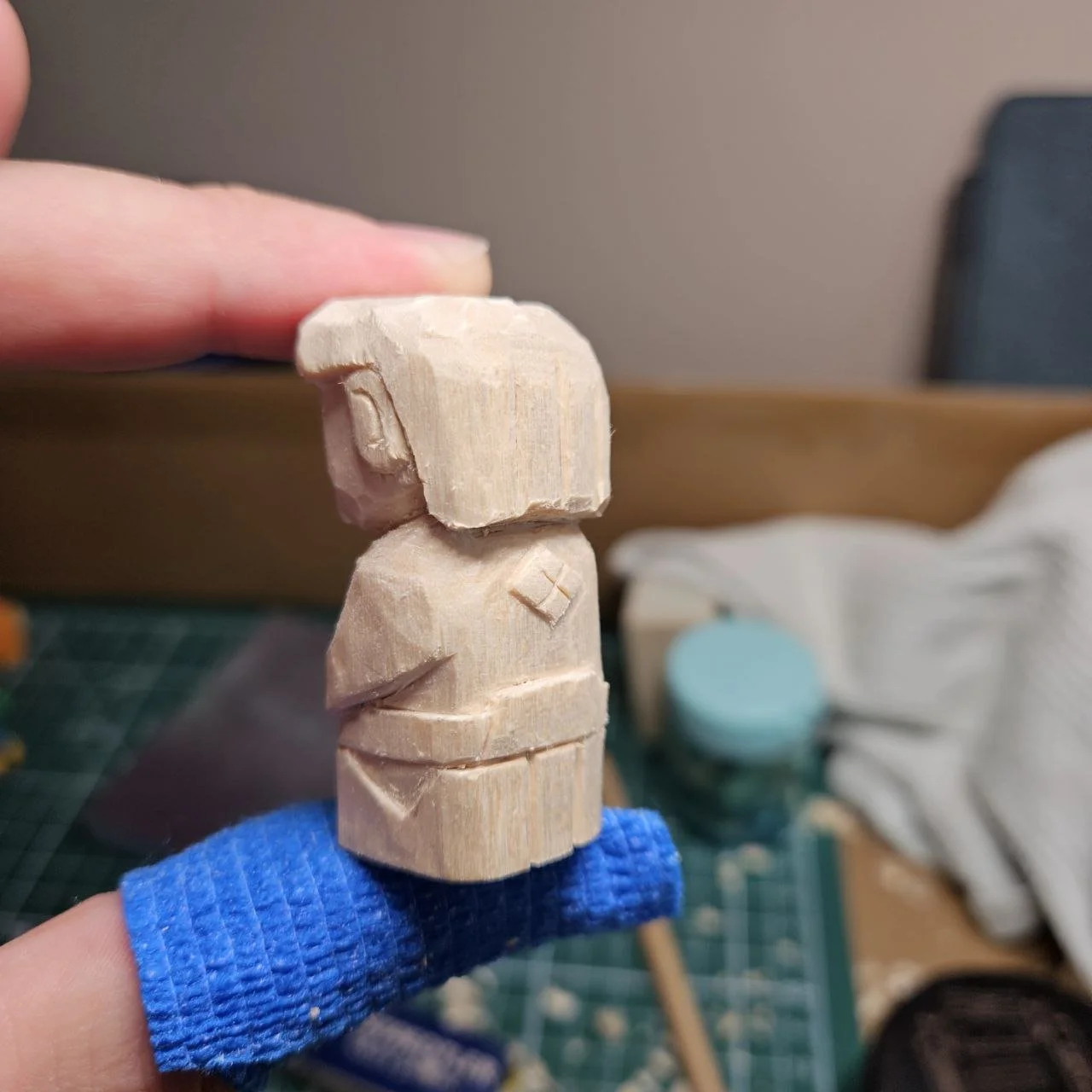 Scultura in legno di una figura umana stilizzata senza dettagli facciali, indossa una maglietta e pantaloni, con i bracci incrociati.