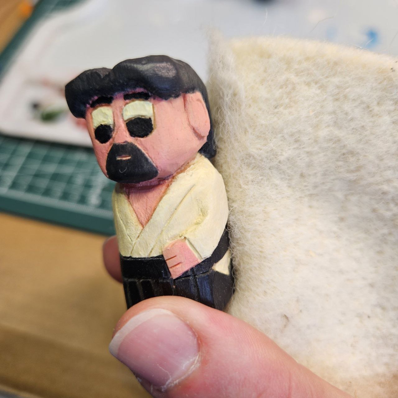 Figura di cartone o di legno di un personaggio con capelli neri, occhiali e barba, vestito con una giacca gialla e pantaloni neri, che tiene un braccio appoggiato a un oggetto bianco e soffice.