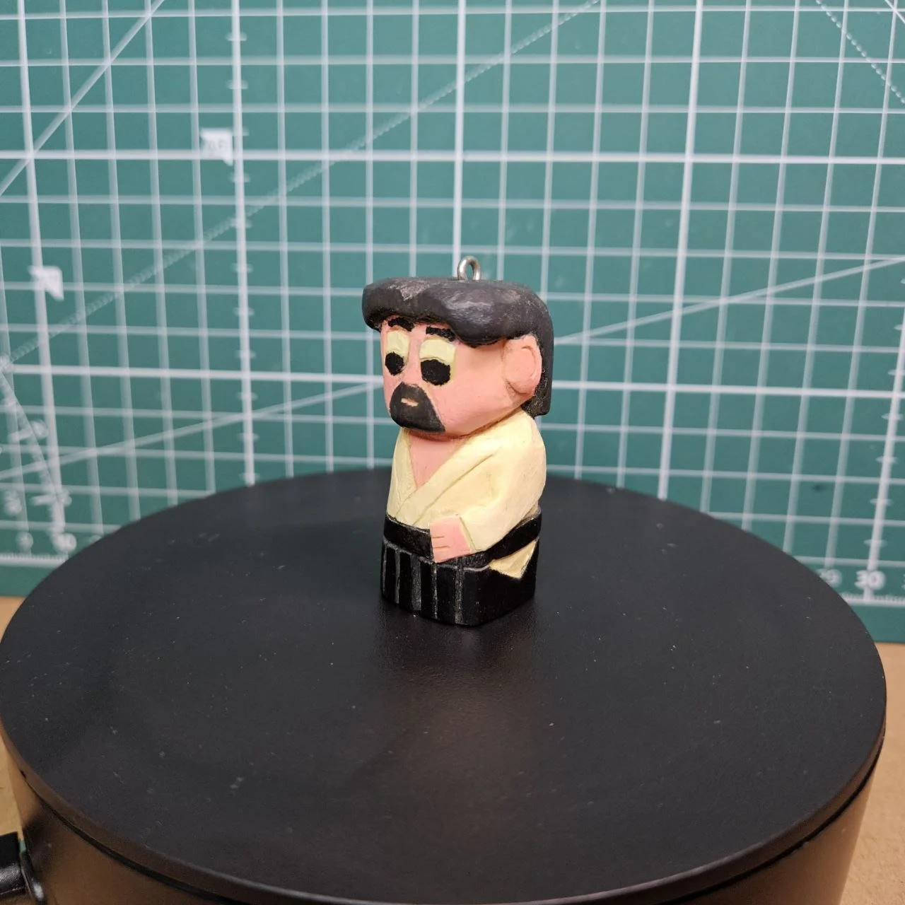 Piccolissima statua in legno o creta di un uomo con barba, capelli neri, occhiali neri, vestito giallo e nero, posizionato su una superficie nera, sfondo con una griglia di taglio e misura