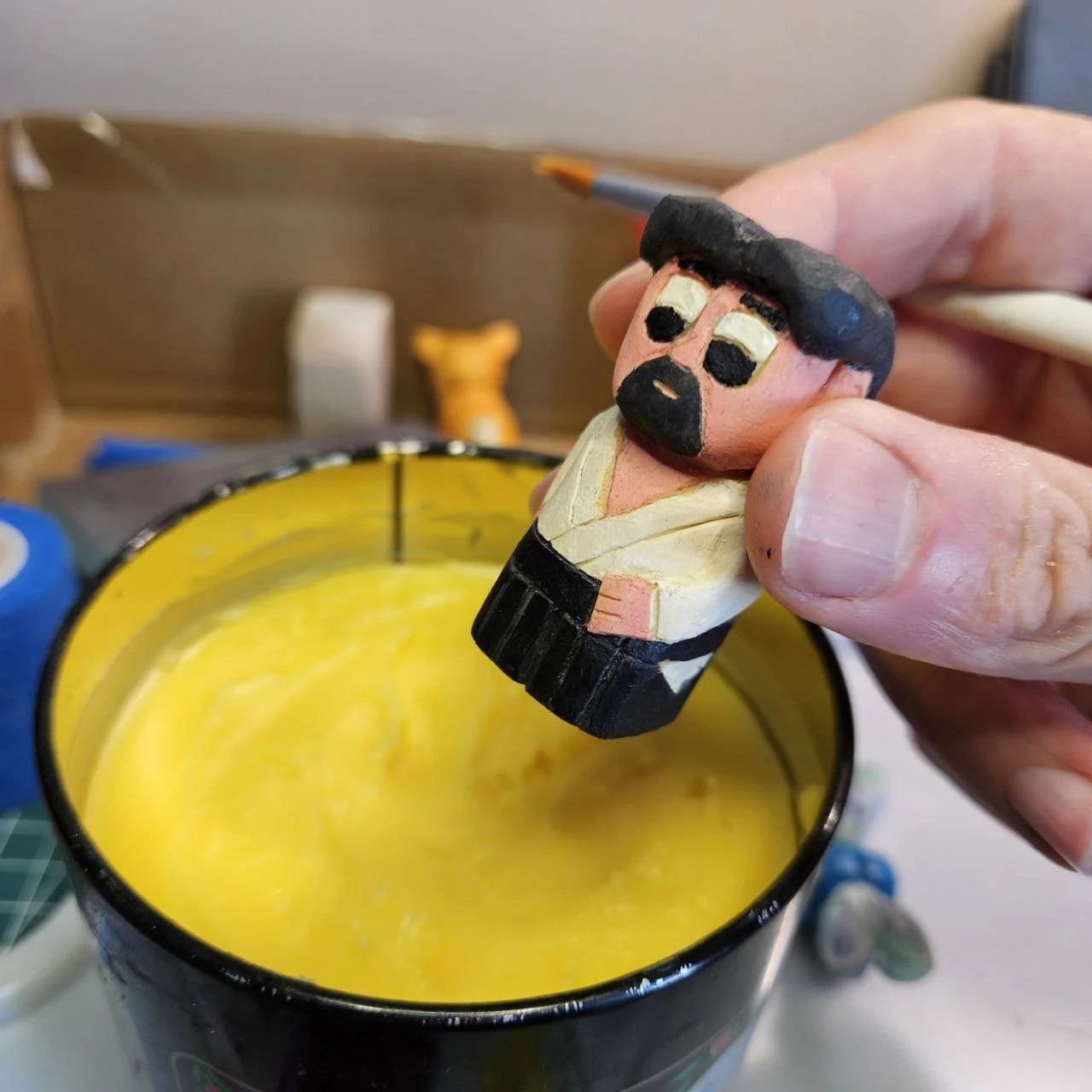 Una mano tiene in mano un piccolo personaggio di legno dipinto che assomiglia a un artista marziale di arti marziali. Sopra una ciotola con una candela gialla. Sullo sfondo, ci sono altri oggetti e un'area di lavoro.