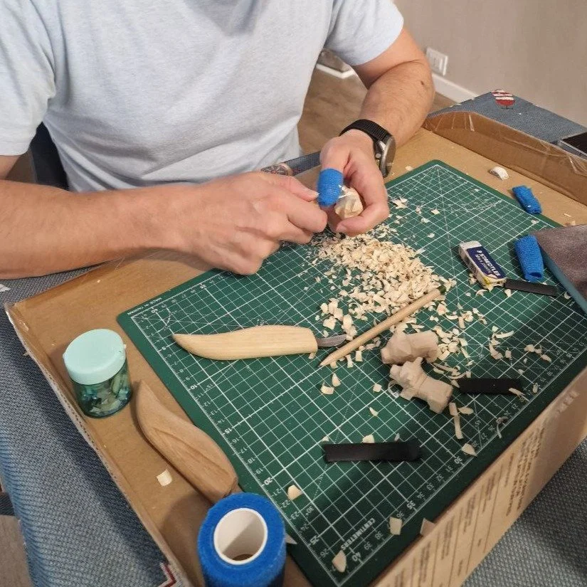 Persona intagliando un oggetto di legno su un tappetino verde con utensili e trucioli di legno sparsi sul tavolo.