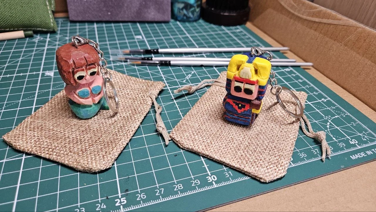 Due portachiavi con figure di legno dipinte, uno con capelli ruggine e occhiali blu, l'altro con capelli gialli, vestito colorato e occhiali, posati su pezzi di tessuto su un tappetino da taglio.