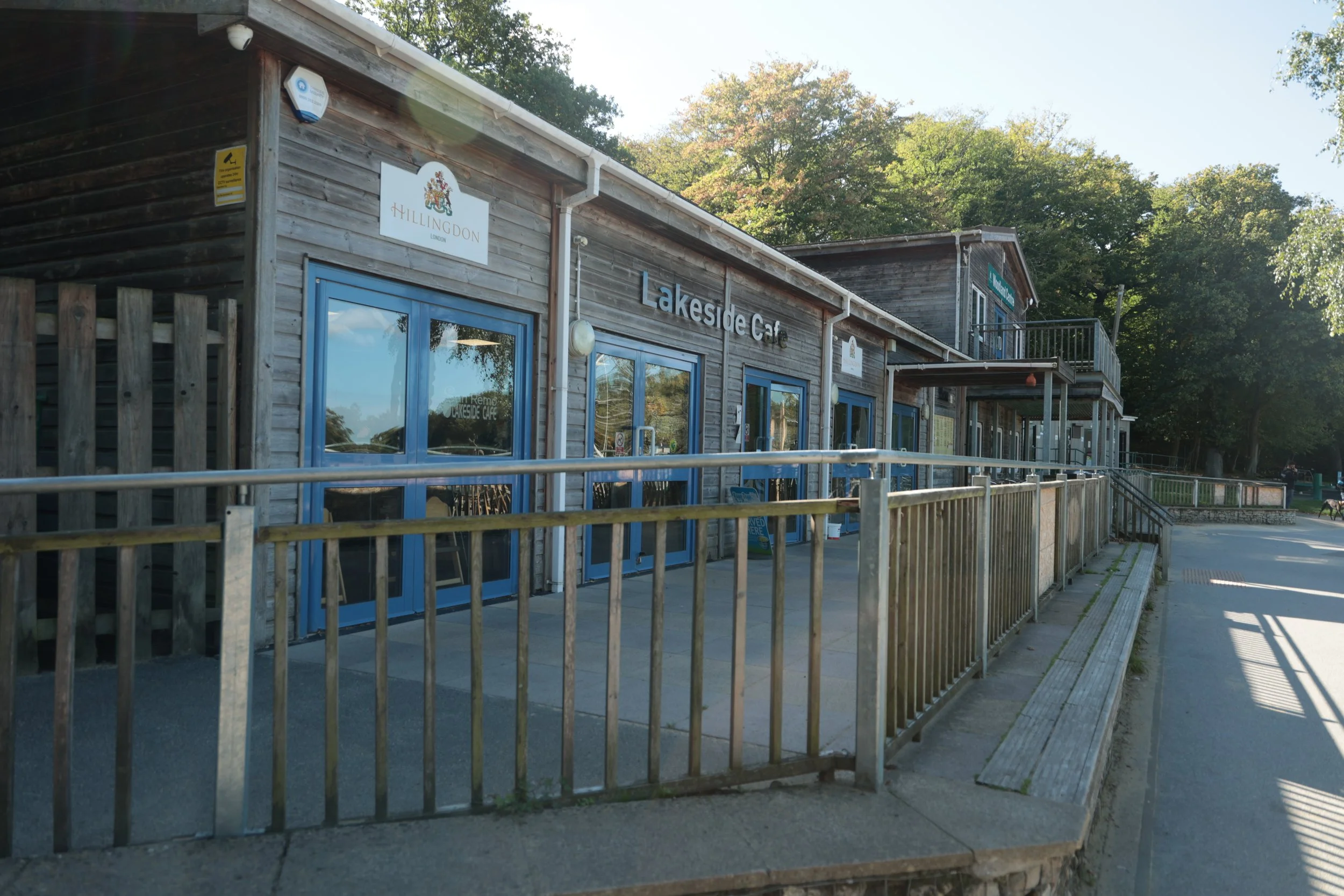 Exterior of Lakeside Cafe housed in a wooden building with blue door frames, surrounded by green trees and a paved outdoor seating area with a metal railing.