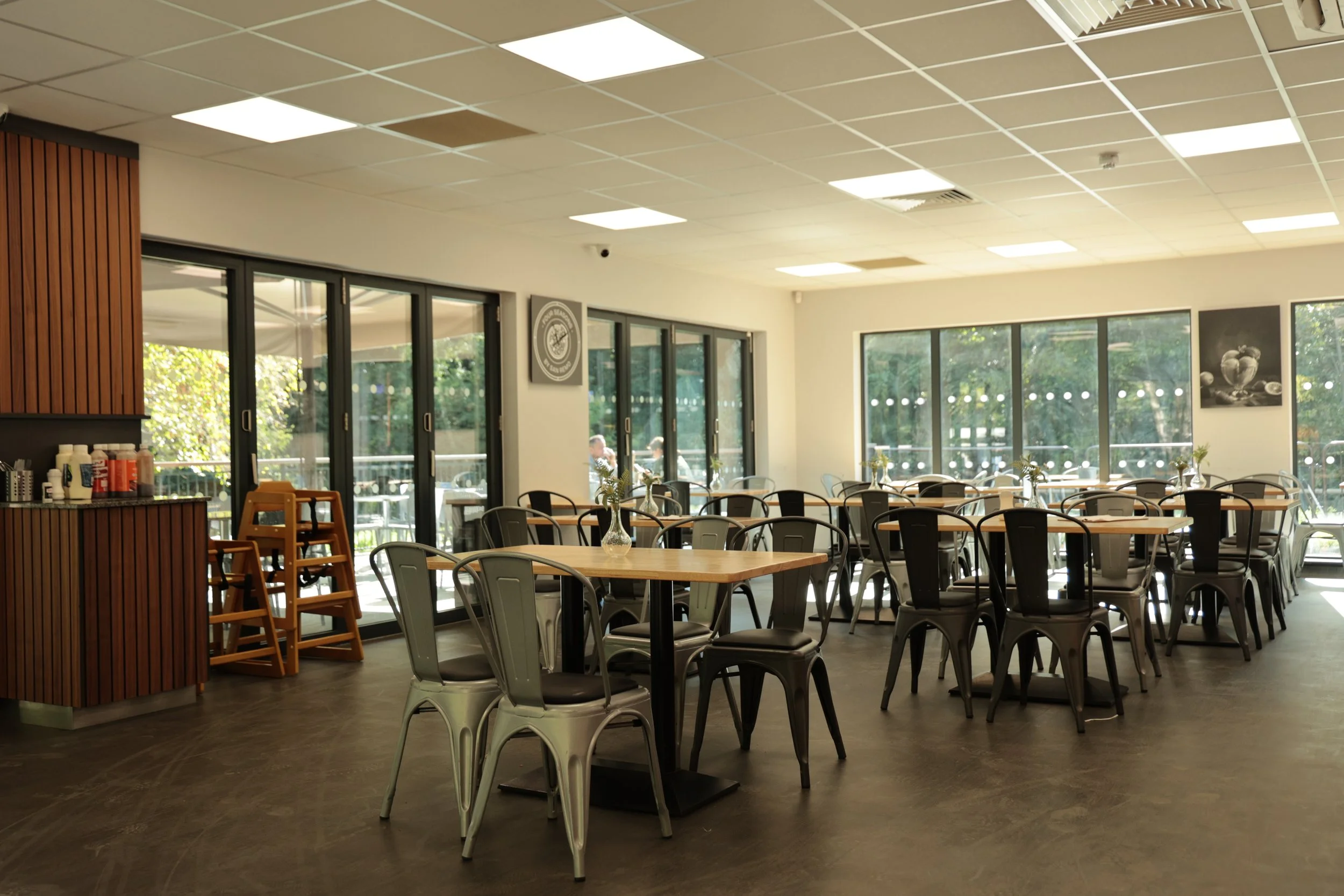 Empty dining area with tables and metal chairs, large windows letting in natural light, a water pitcher on a table, and wall art, in a modern restaurant or cafe.