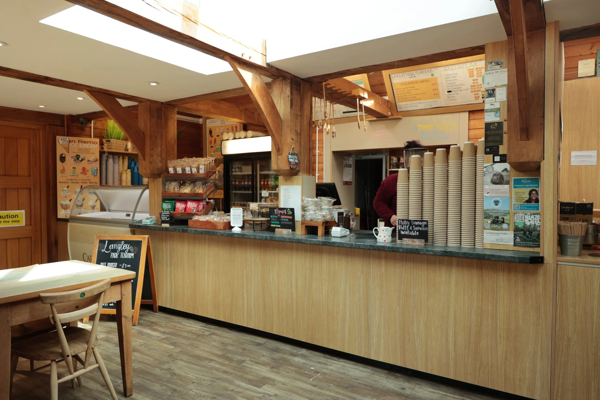 Interior of a cafe or ice cream shop with a wooden counter, stacks of paper cups, and a menu hanging above. A person behind the counter prepares an order, and there are various packaged snacks and ice cream chillers visible.