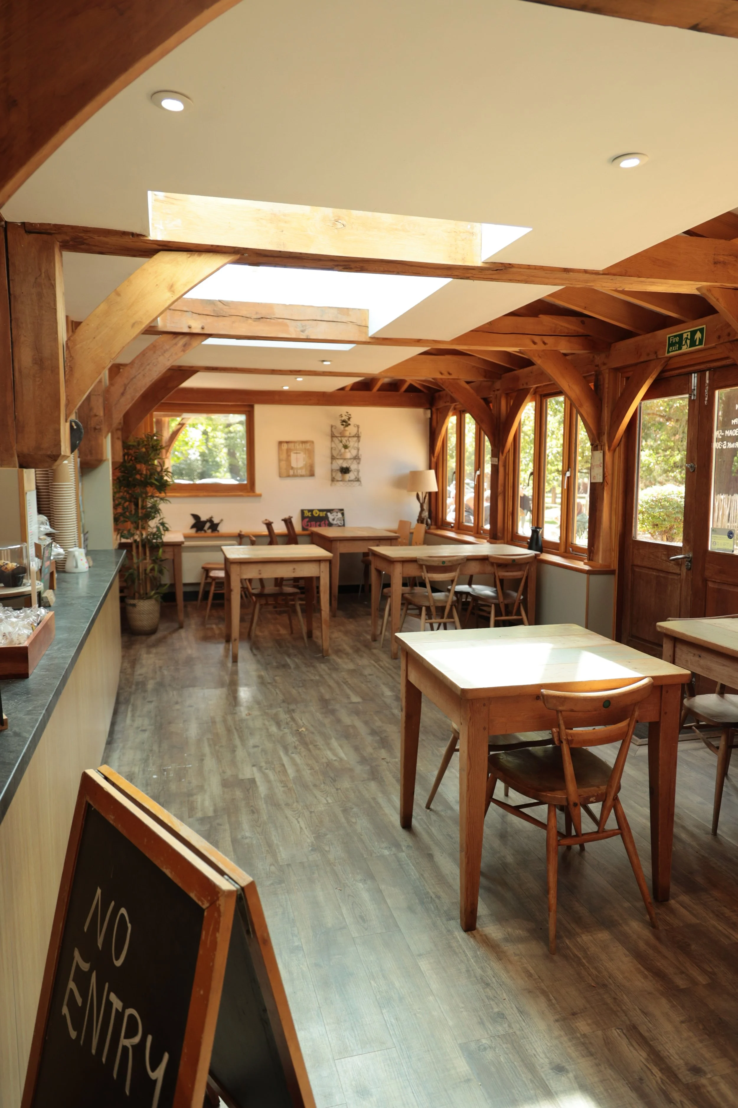 Empty dining area with wooden tables and chairs, sunlight coming through large windows, and a ceiling with wooden beams and skylights.