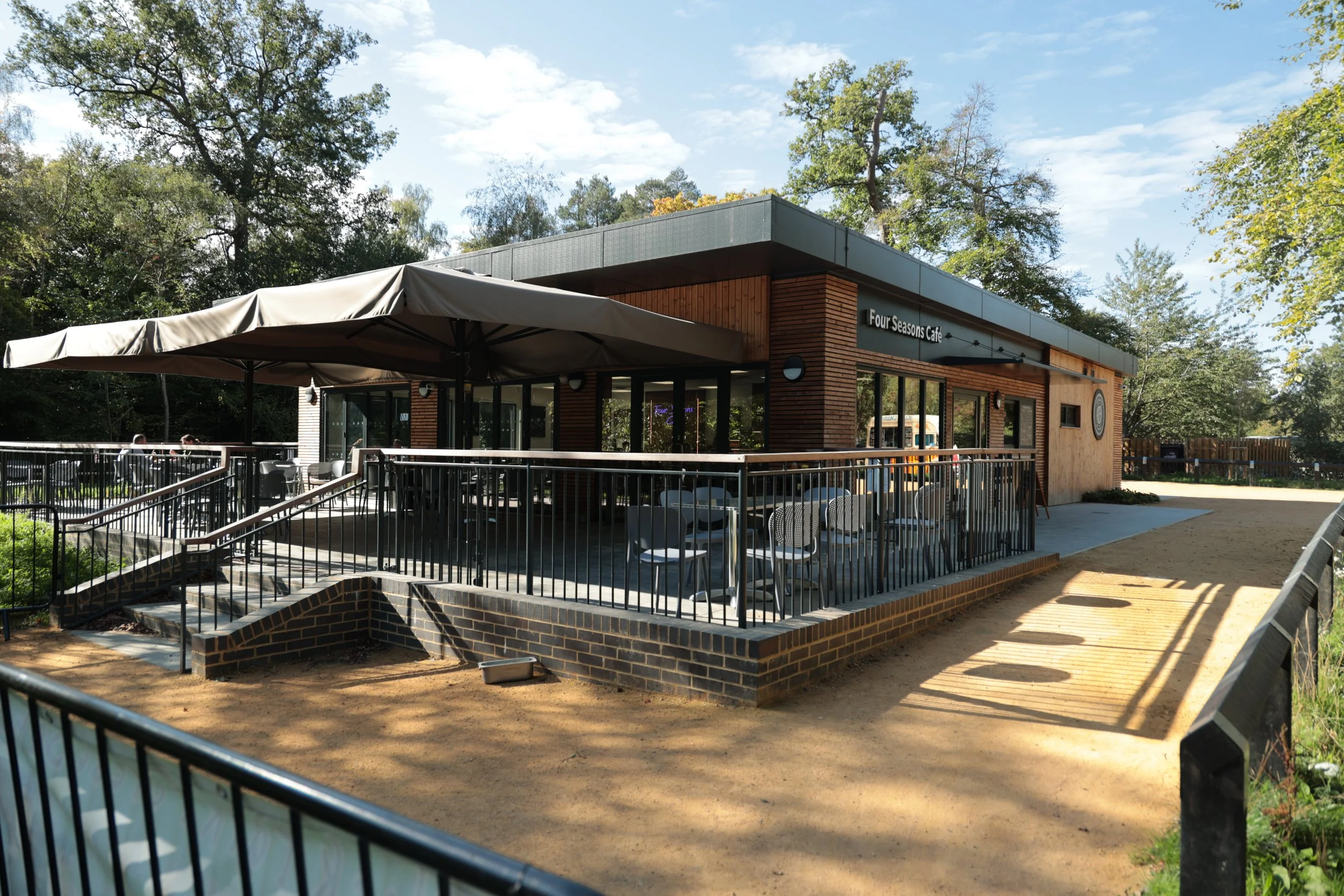 Exterior view of Four Seasons Cafe with outdoor seating area, black railings, large umbrella, and trees in the background.