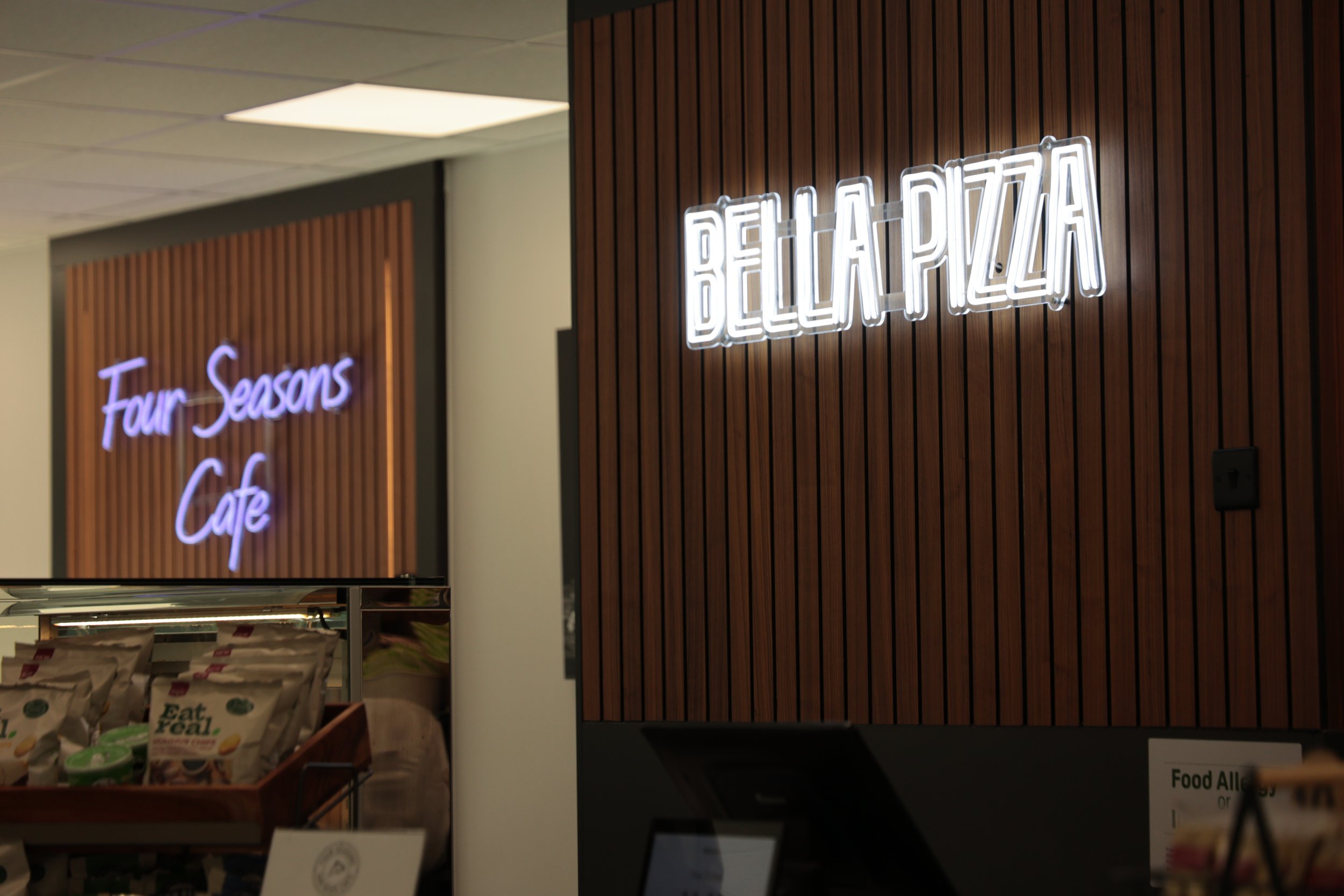 Neon sign reading 'BELLA PIZZA' on a wooden wall, with another sign in the background saying 'Four Seasons Cafe' in purple cursive lights.