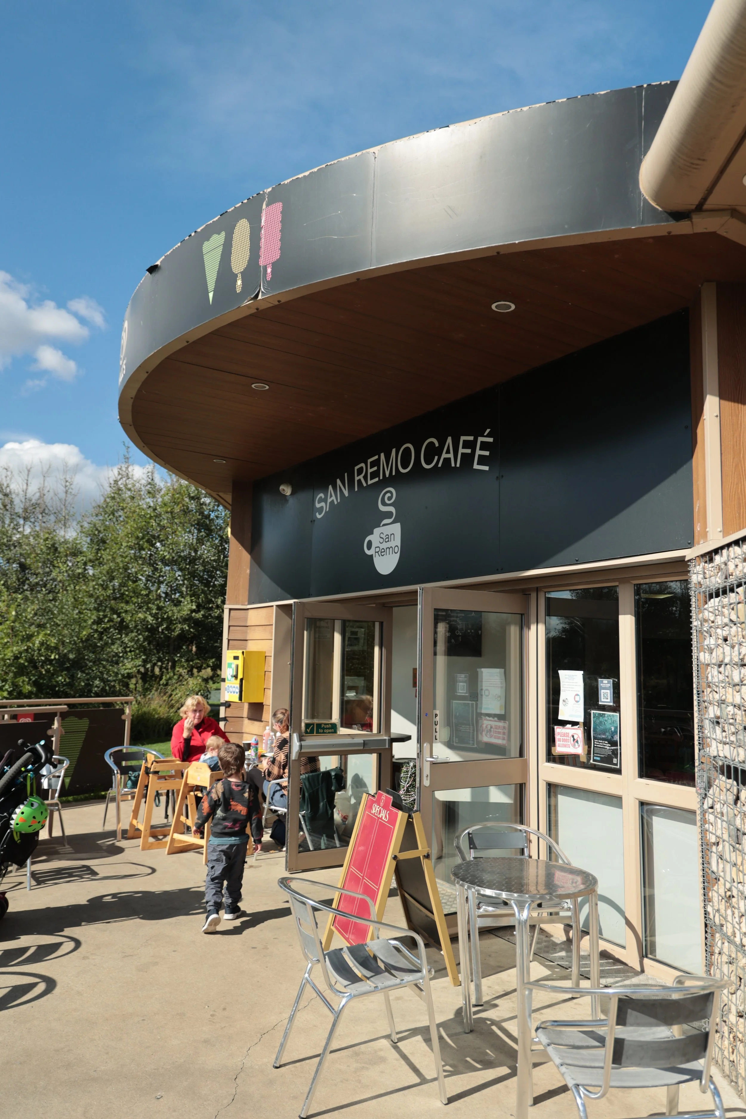 Exterior view of San Remo Café with people sitting outside, children playing, and clear blue sky.