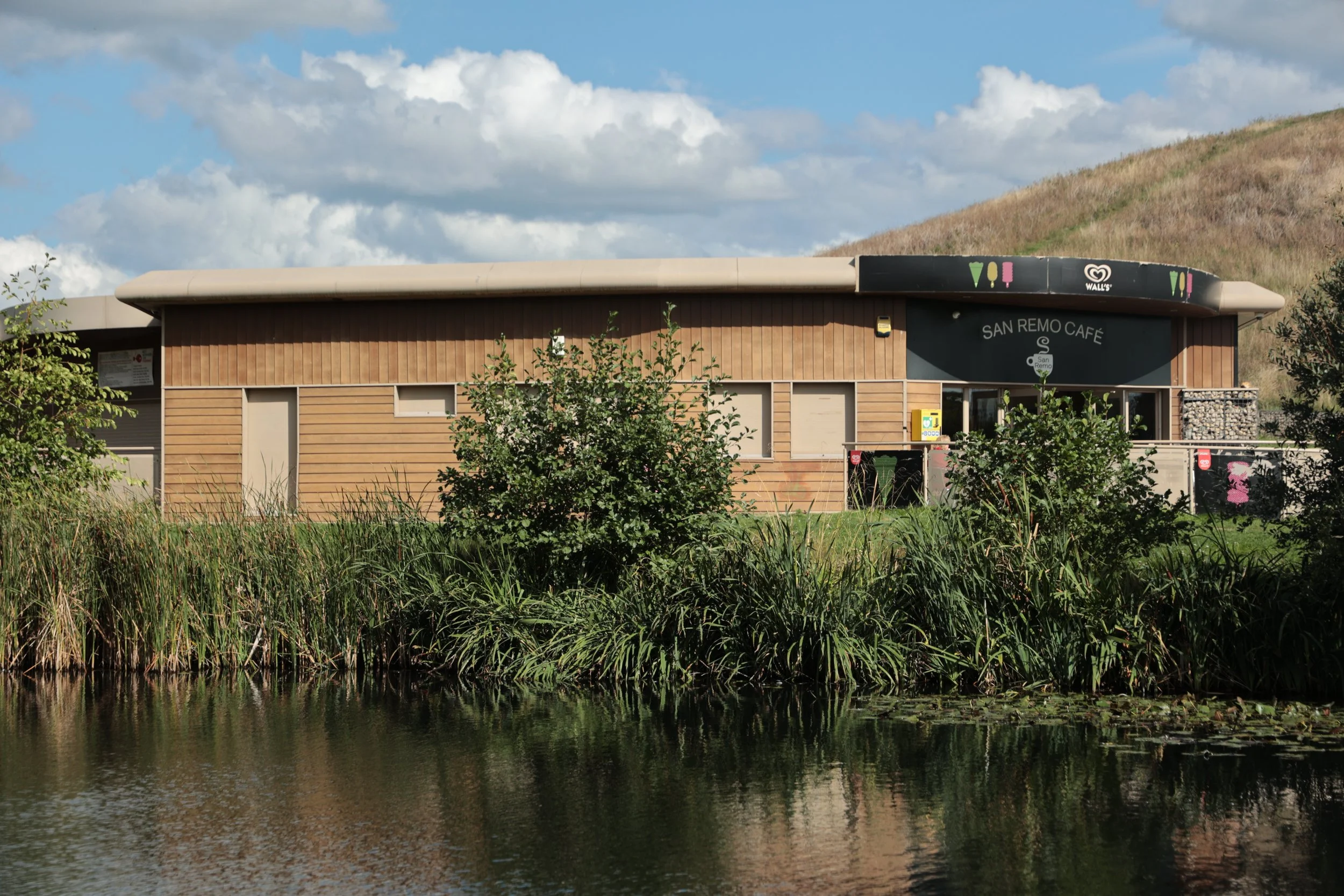 A single-story wooden building with the sign 'San Remo Café' on a curved awning, located near a body of water with tall grass and bushes in front, and a hillside and cloudy sky in the background.