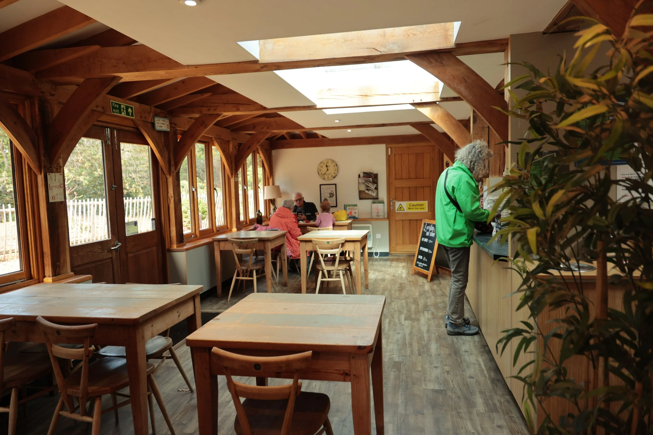 A cozy cafe with wooden furniture and large windows, a woman in a green jacket orders at the counter, a group of people sit at a table in the background, sunlight streams through the skylights.