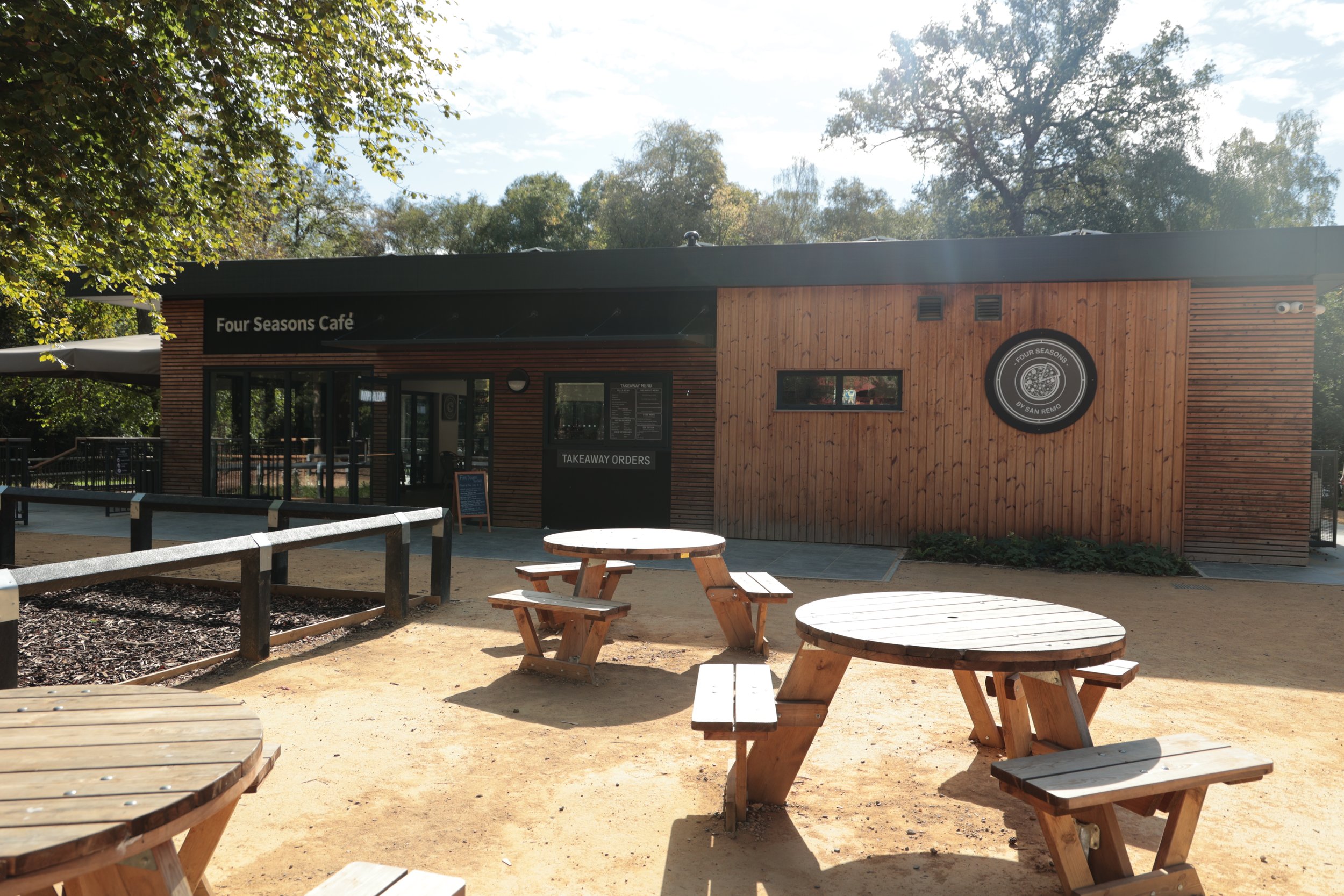 View of a wooden cafe building named 'Four Seasons Cafe' with outdoor seating, including round wooden tables and benches, and trees in the background.