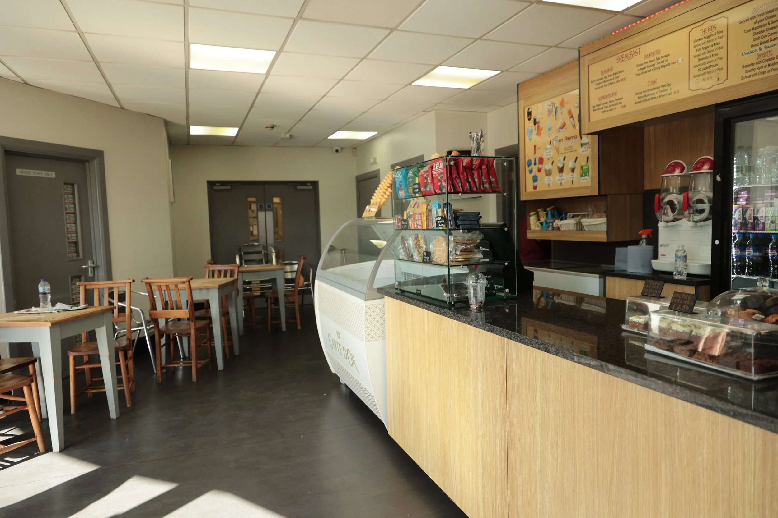 Empty indoor cafe with wooden tables and chairs, checkout counter with snacks and drinks, and a menu on the wall.