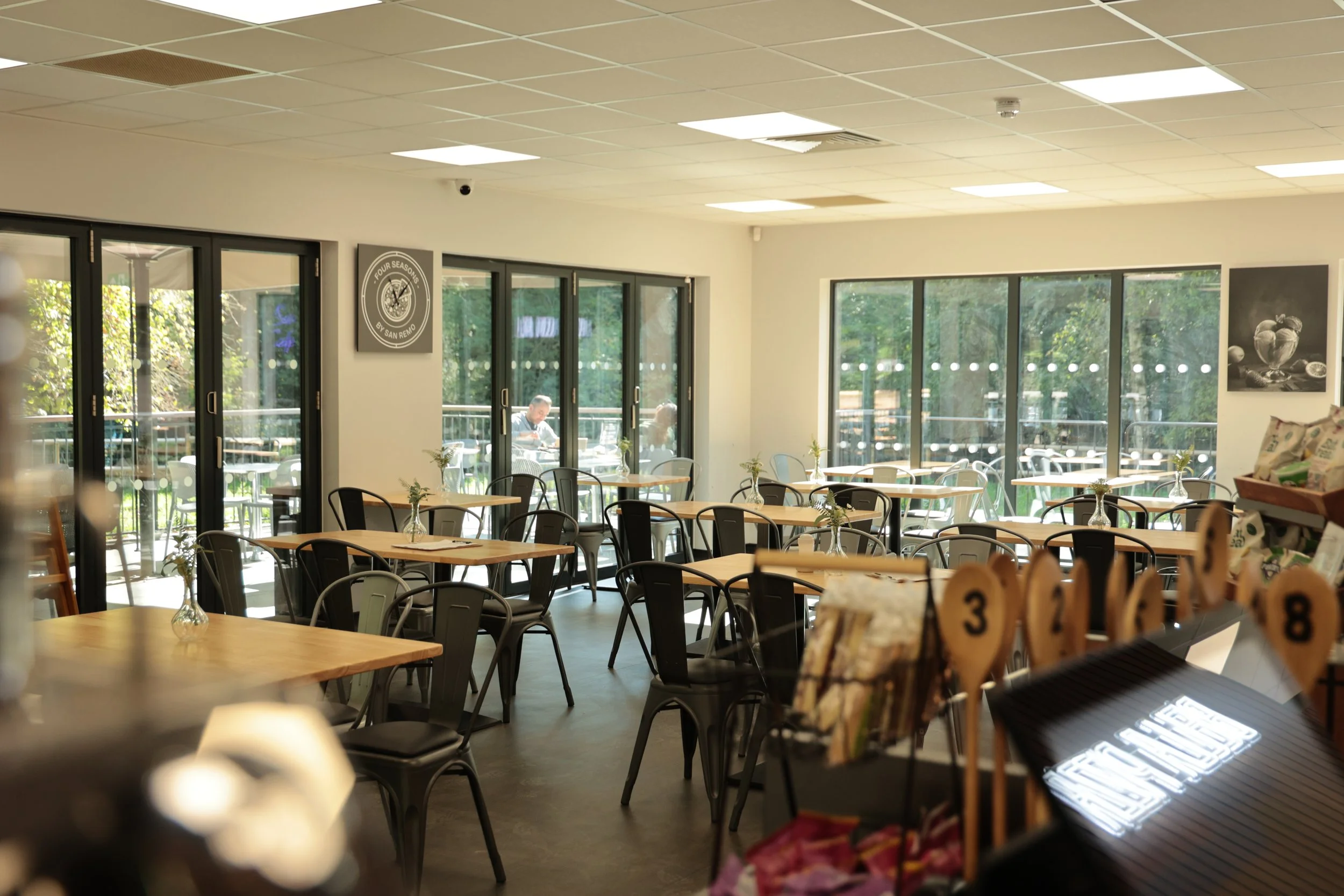 Empty restaurant with wooden tables, black chairs, and large windows with outdoor seating