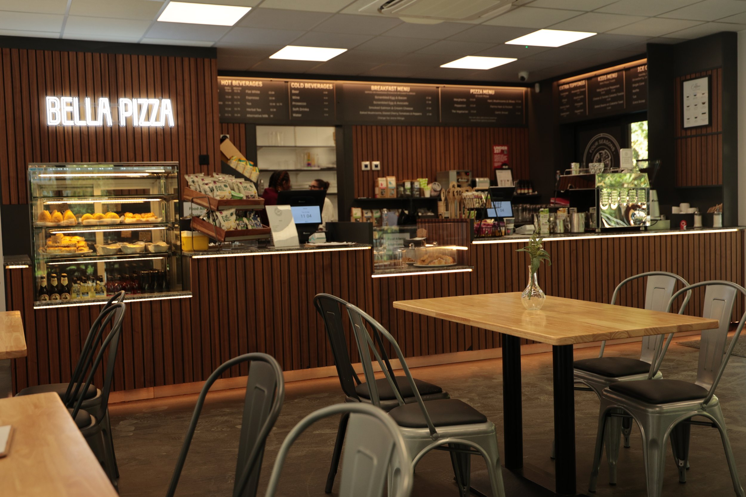 Interior of restaurant with a wooden counter, menu boards, and seating area with wooden tables and metal chairs, one table has a vase with flowers.