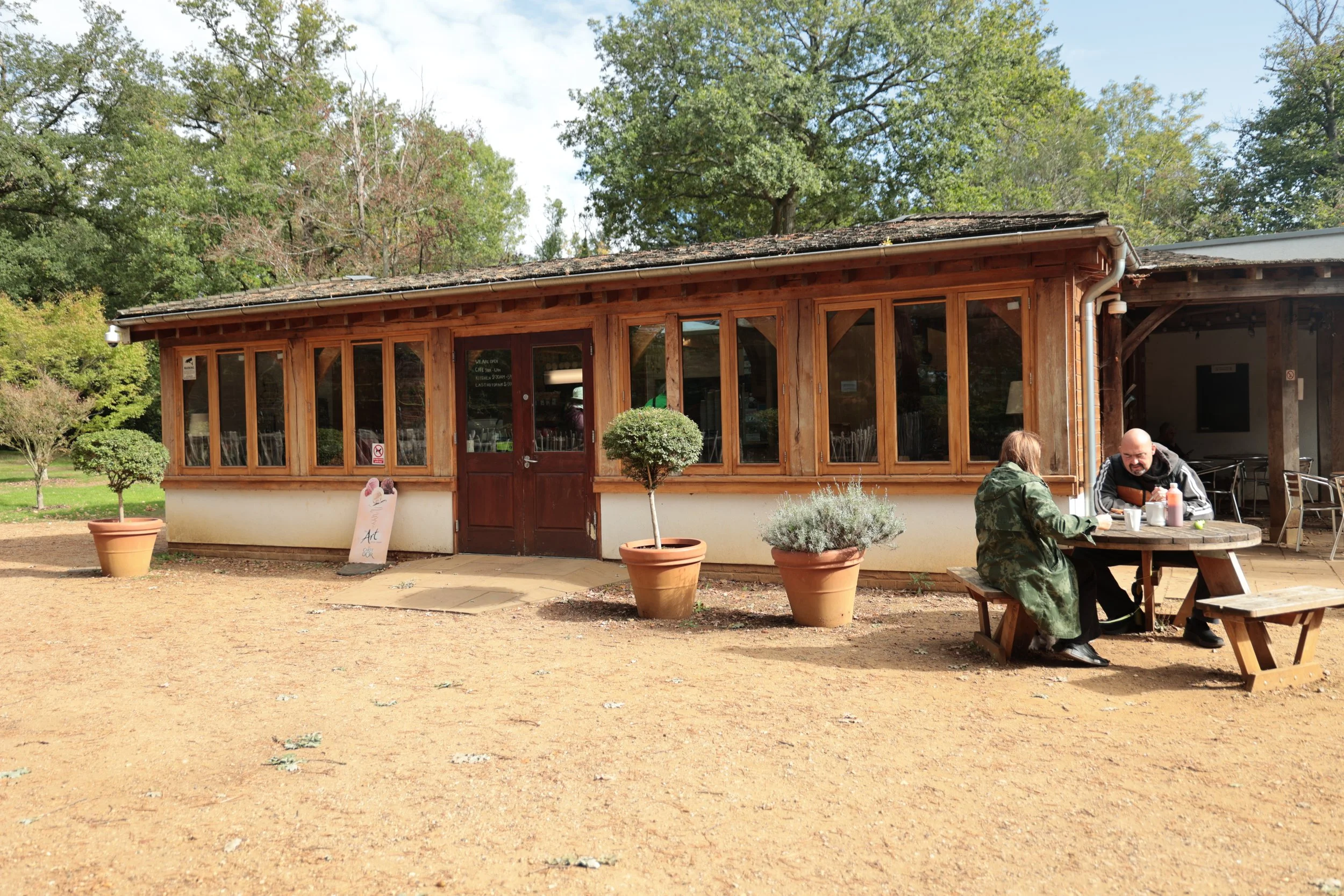 A small rustic café with wooden exterior and large windows, two people sit at a picnic table outside, trees in the background, potted plants around the building.