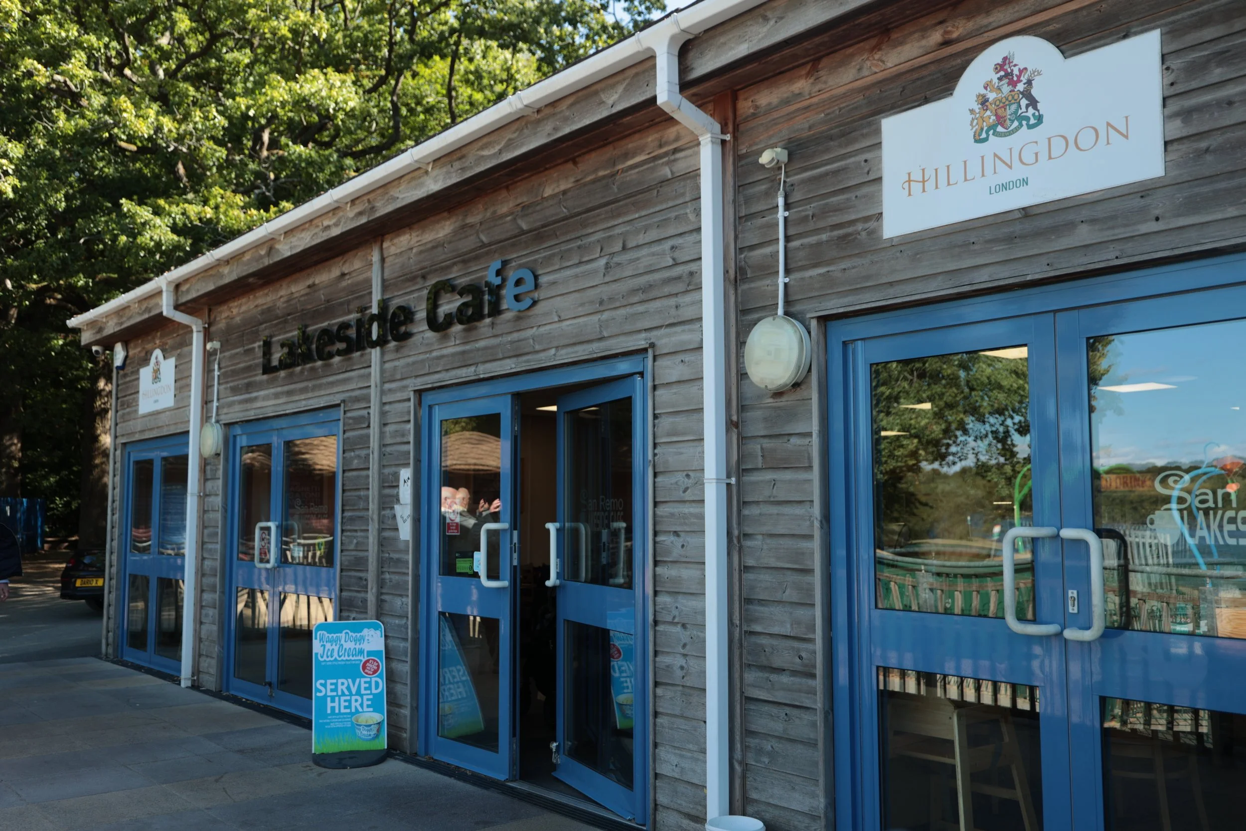 Exterior of Lakeside Cafe, a wooden building with blue window and door frames, signage with the name Hillington London, and a small sign indicating ice cream is served here. Trees and parked cars are visible in the background.