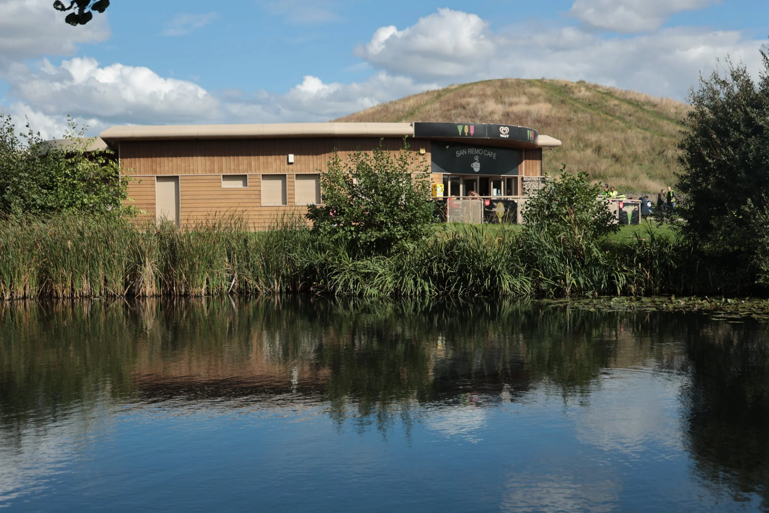 A building labeled 'San Remo Café' with a wooden exterior, situated near a body of water with reflections. The background includes a grassy hill and a partly cloudy sky. There are some people visible on the right side of the building.