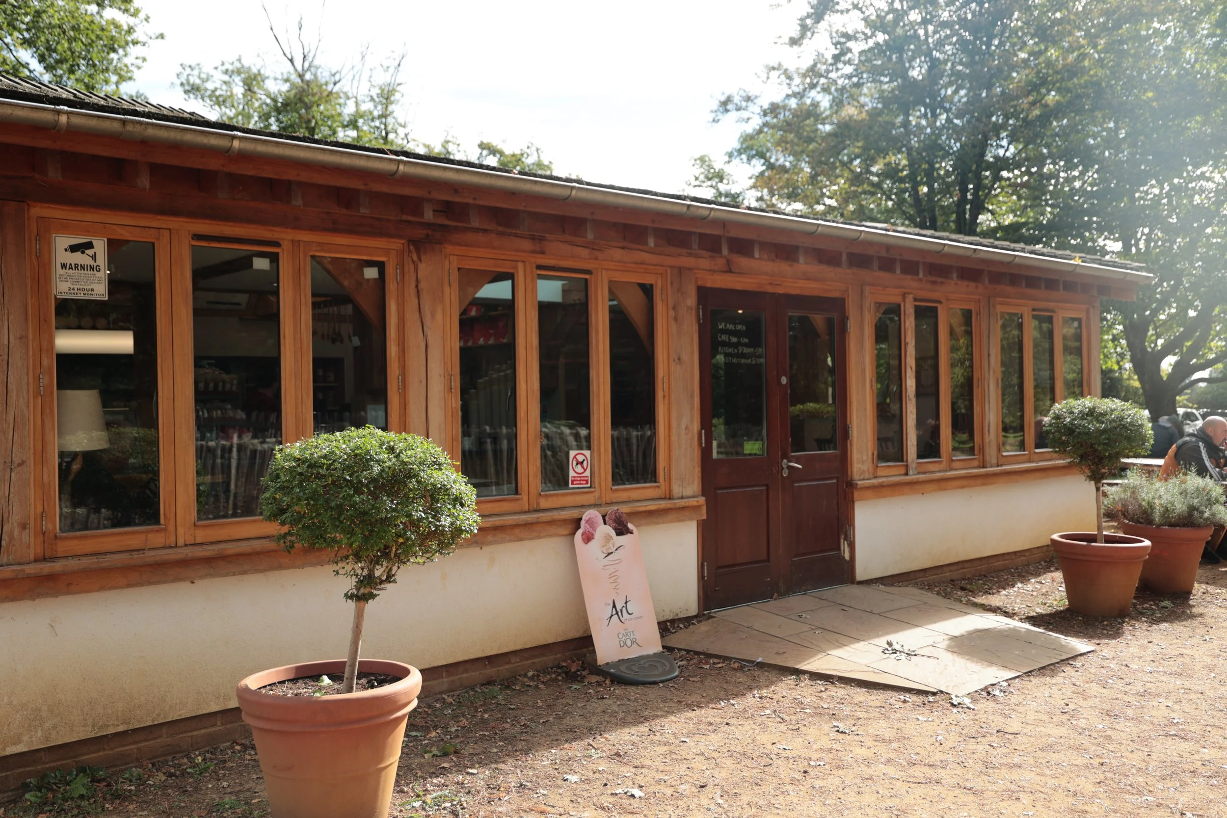 Exterior of a wooden building with large window panes and a closed wooden door, potted plants in front, and an art signboard nearby, set on a dirt ground with trees in the background.