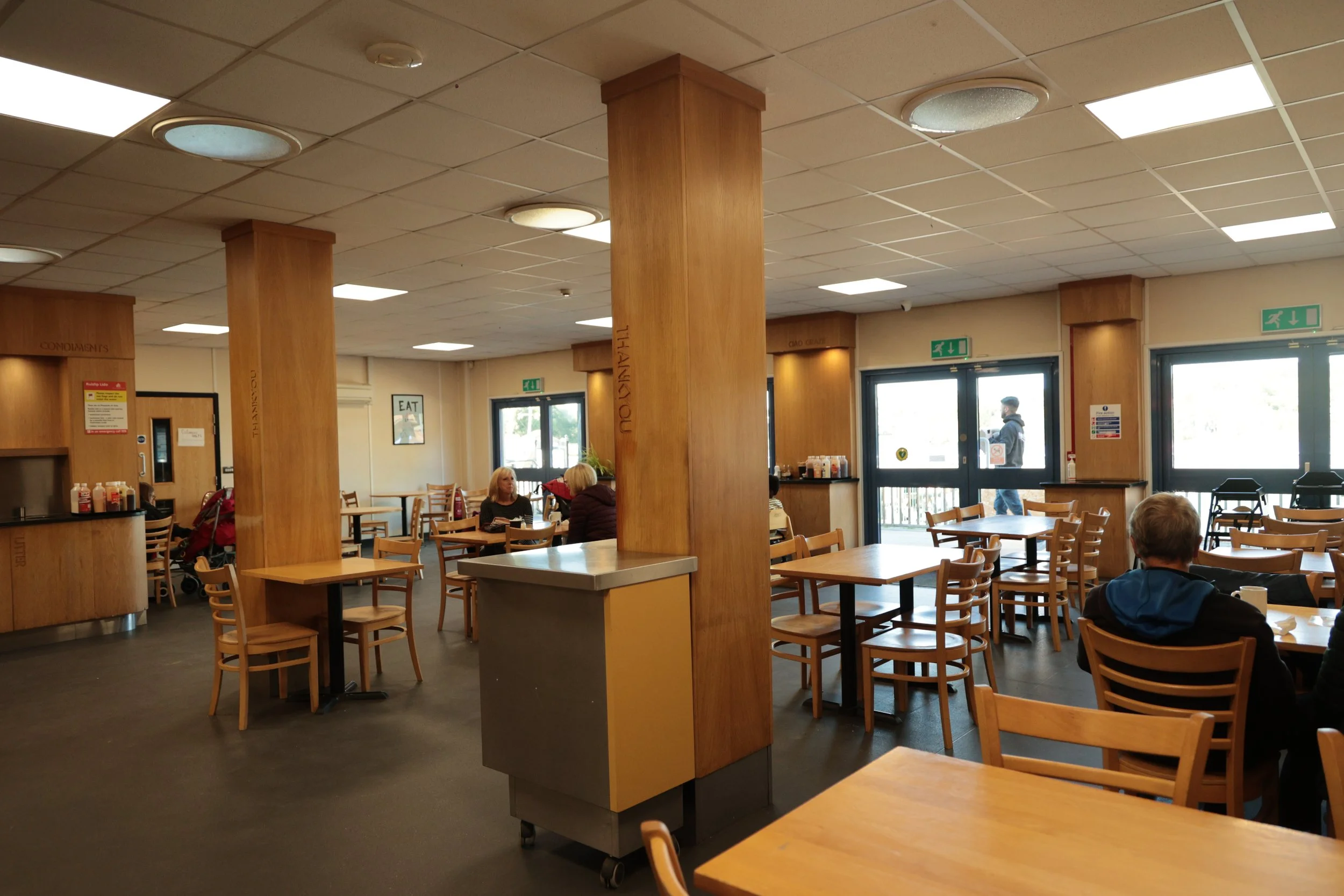Interior of a casual restaurant with wooden tables and chairs, a large wooden support column in the center, and windows letting in natural light. A few customers are seated, and a man is walking outside through the glass door.