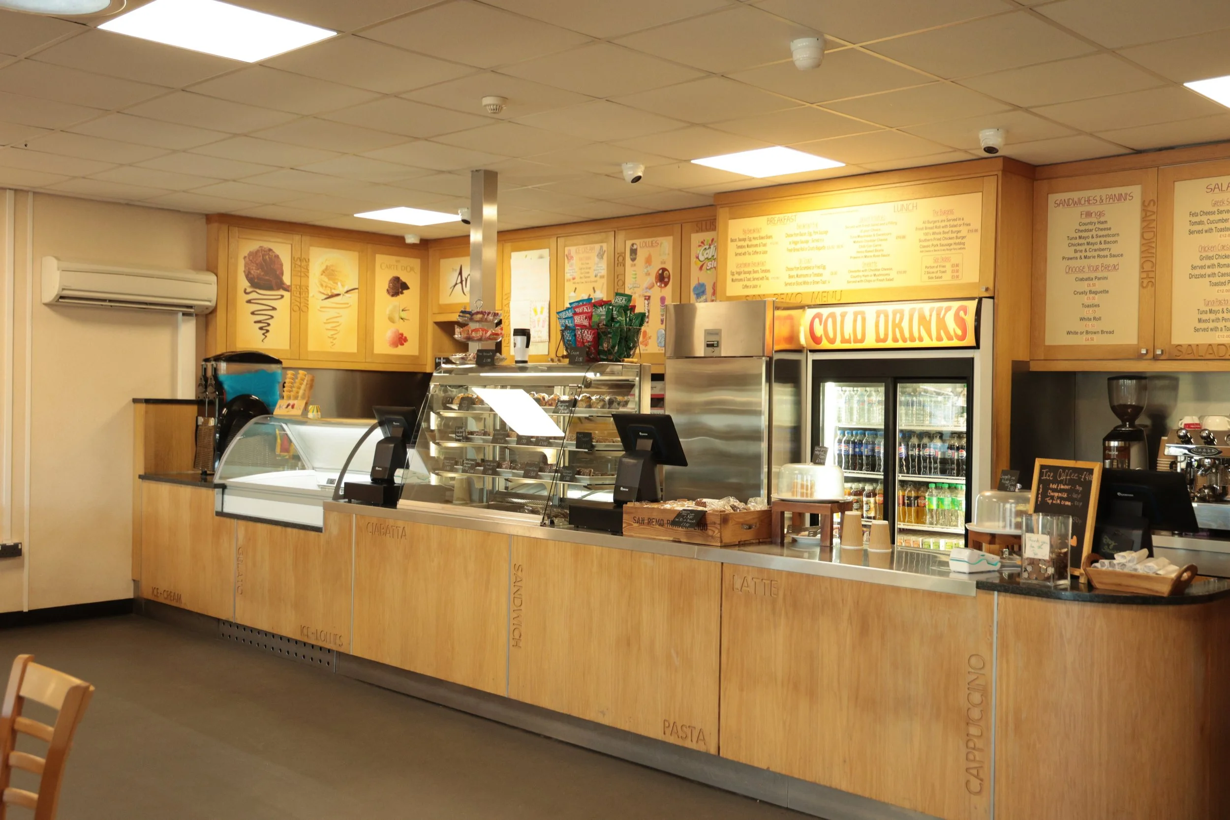 Inside a fast-food cafe with a wooden counter, a refrigerated display case, and a large sign saying 'Cold Drinks.' Menu boards and different snacks are visible behind the counter, along with a coffee machine and a coffee grinder.