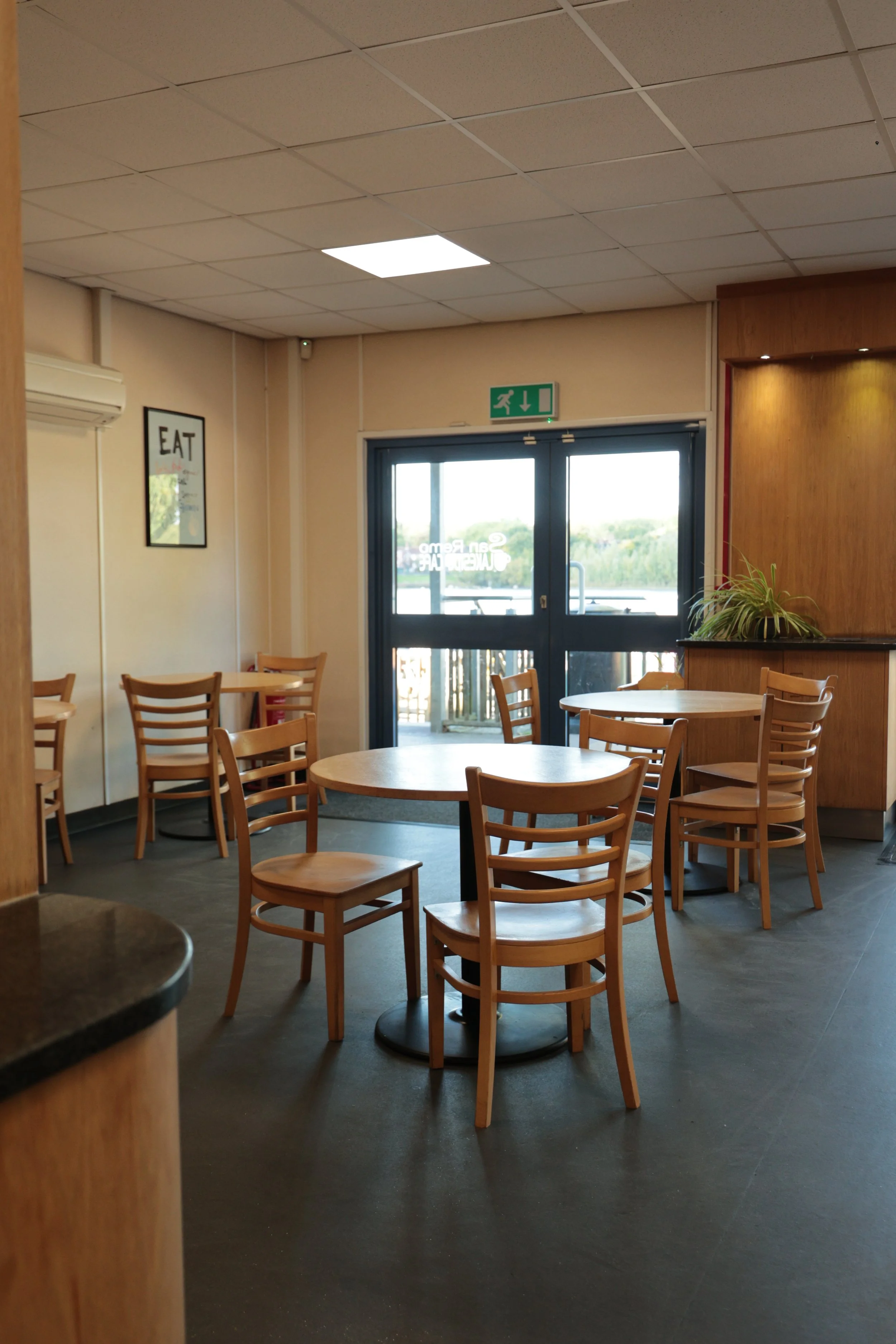 An empty cafe or restaurant with wooden chairs and tables, a door leading outside, and a plant on a counter, with a small sign on the wall that says "EAT."