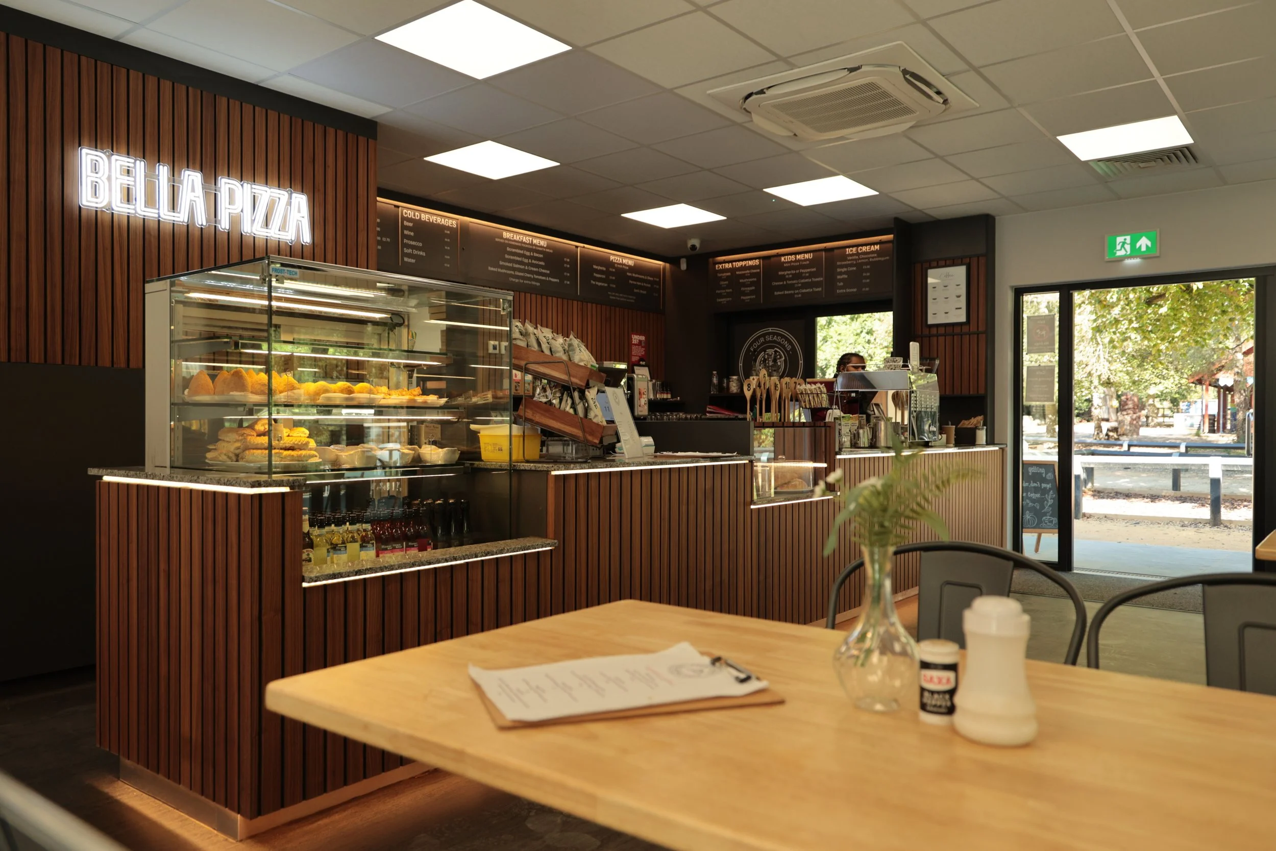 Interior of a modern pizza restaurant with a wooden counter, glass display case with sandwiches, a menu hanging on the wall, and a table with a menu and napkins in the foreground. Outside is visible through the door, showing trees and a sunny day.