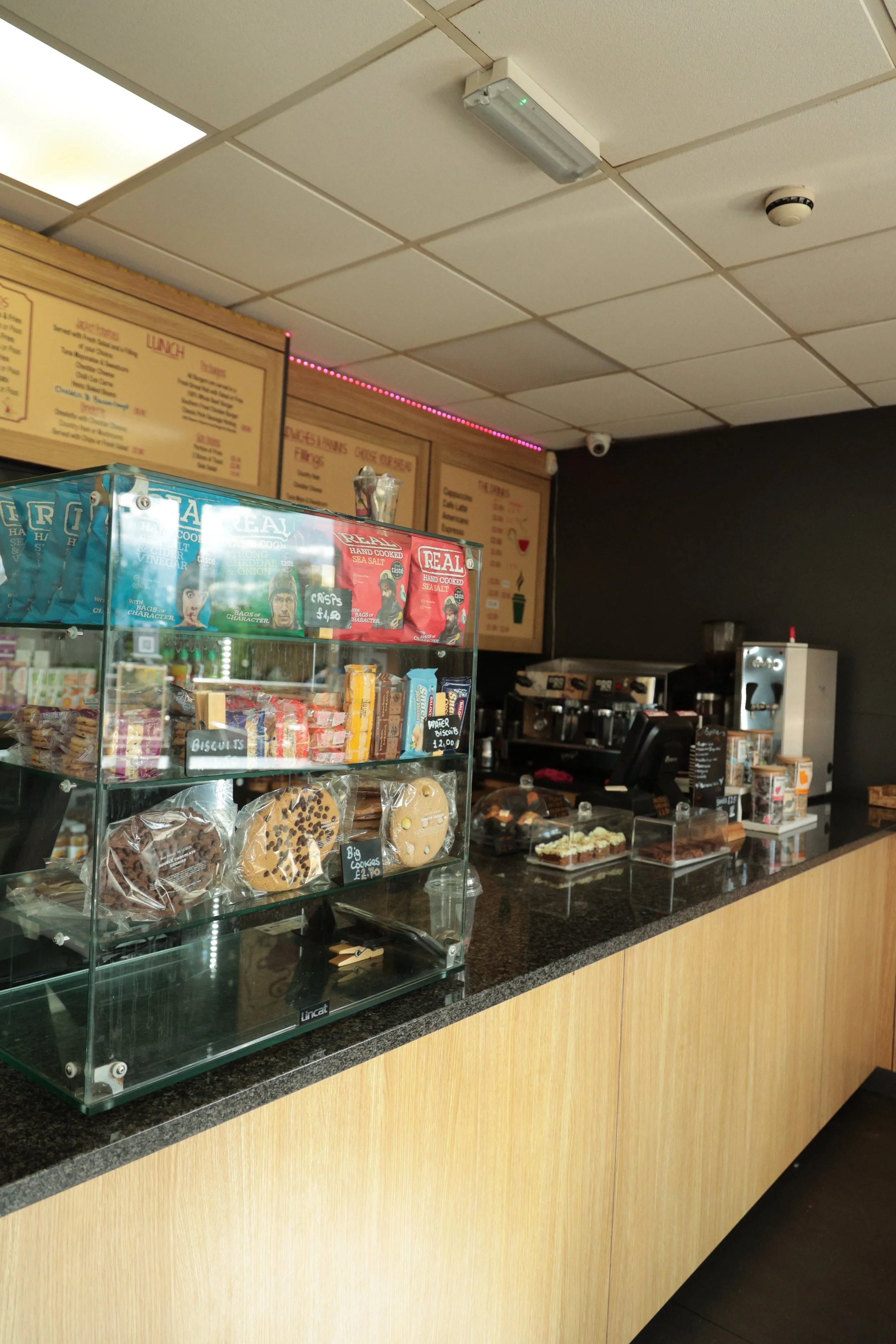 Corner of a cafe counter with baked goods, snacks, and a coffee machine, with menu boards on the wall above.