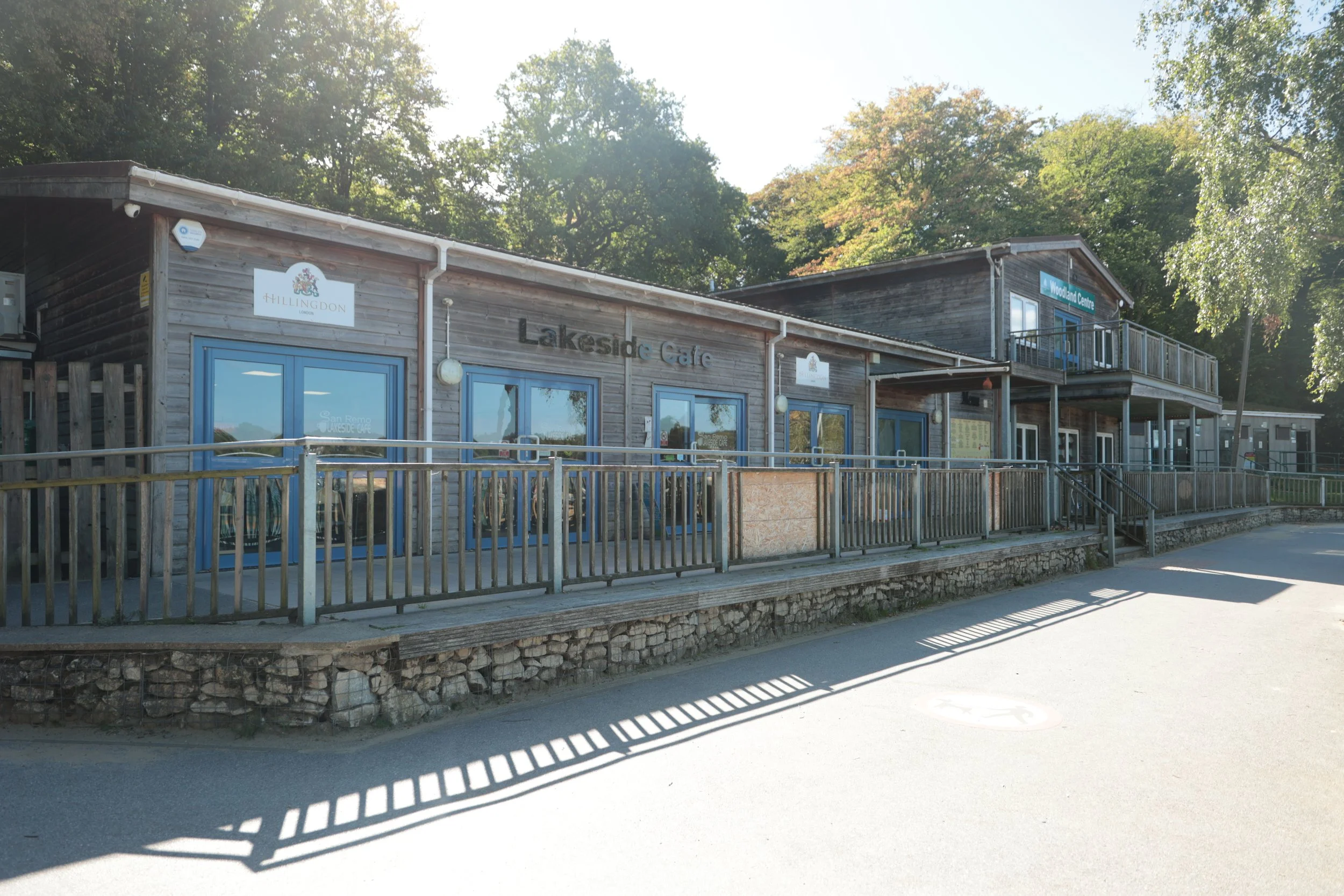 A wooden building with blue doors and windows, labeled 'Lakeside Cafe,' with signs for 'Hillingdon' and 'Woodland Centre,' surrounded by trees, with a ramp and stairs leading to the entrance, and a paved area in front.