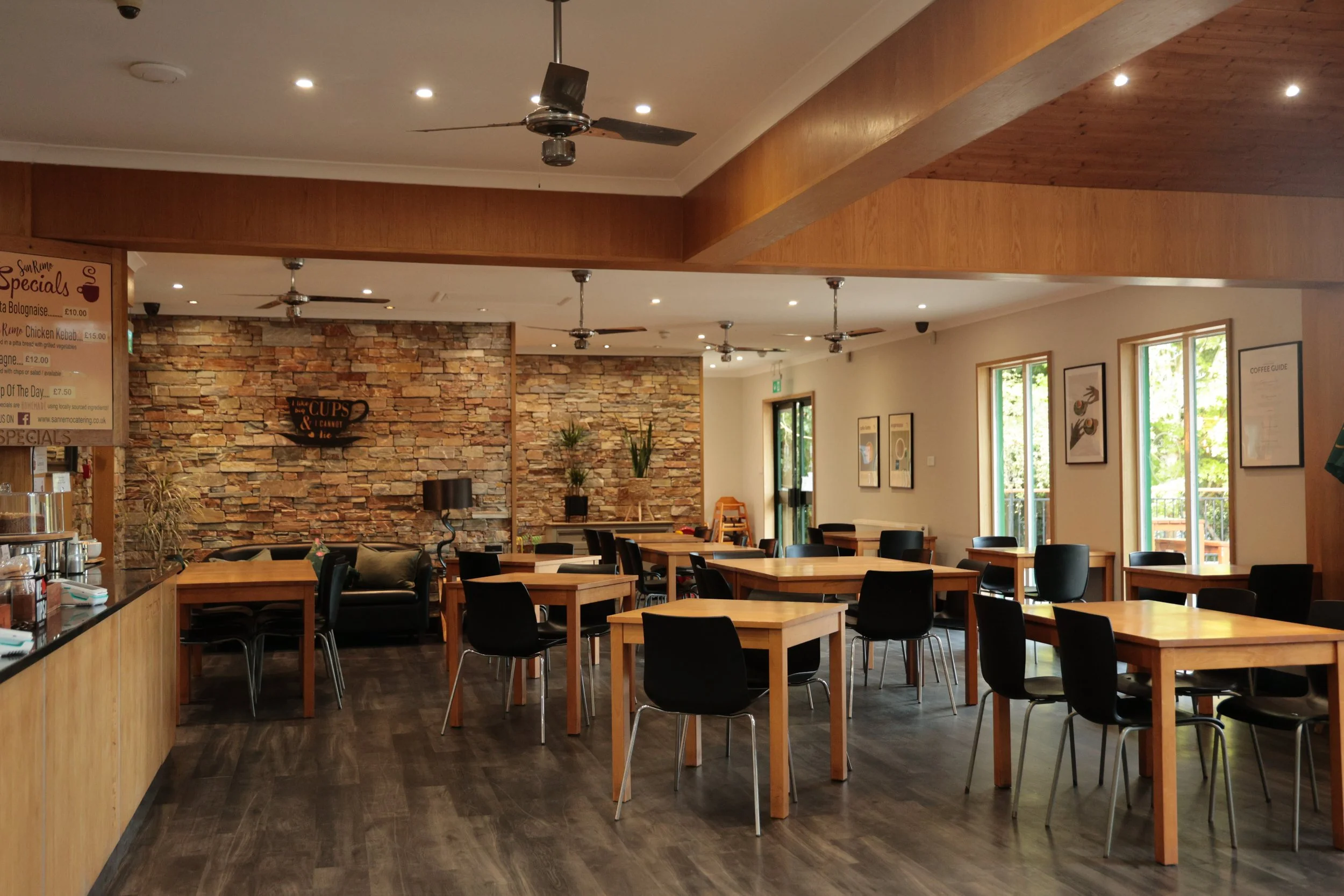 Interior of cafe with wooden tables and black chairs, decorated with framed pictures on the walls, large windows letting in natural light, and potted plants