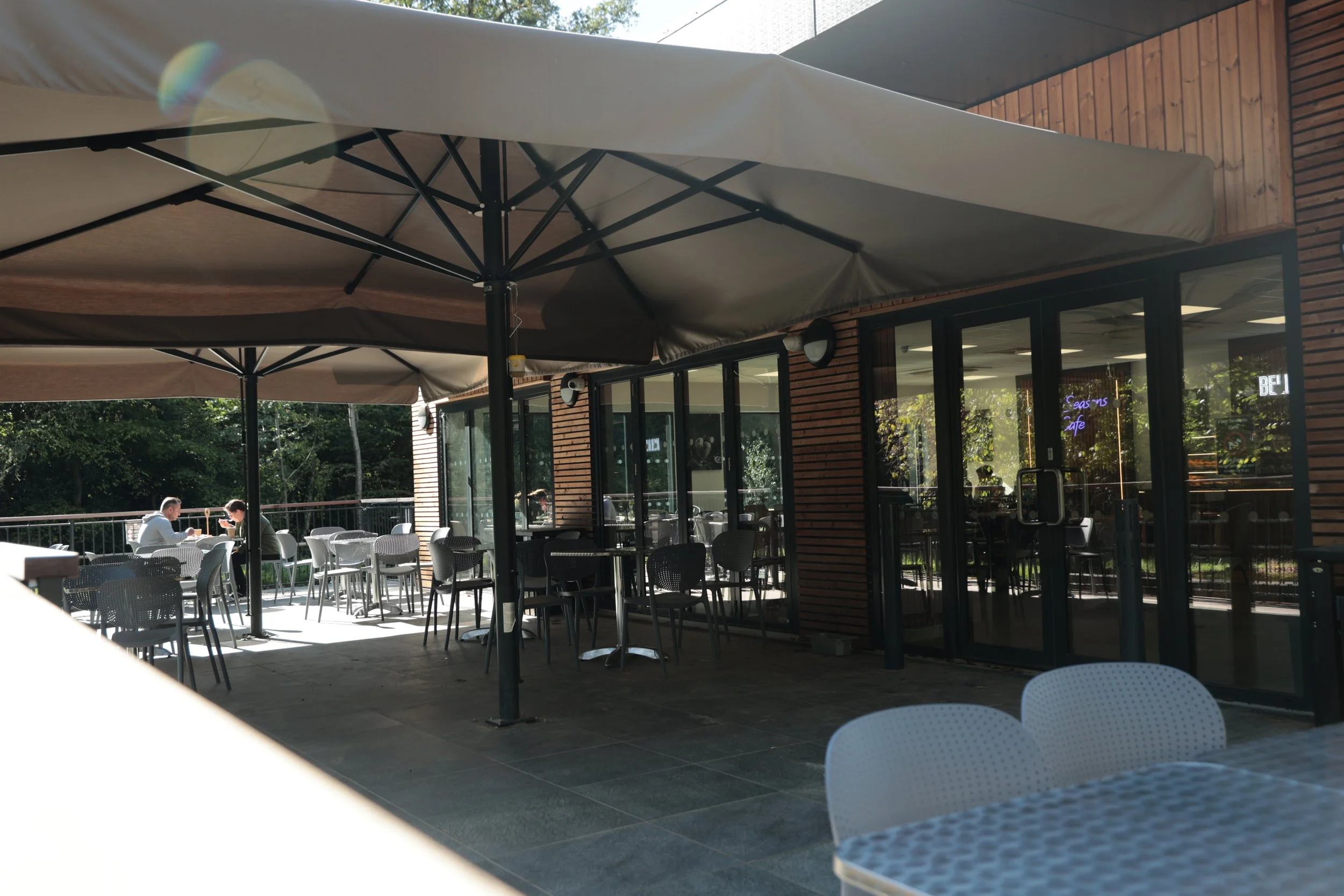 Outdoor patio of a cafe or restaurant with tables and chairs, large umbrellas for shade, and a glass building facade reflecting trees and sky.