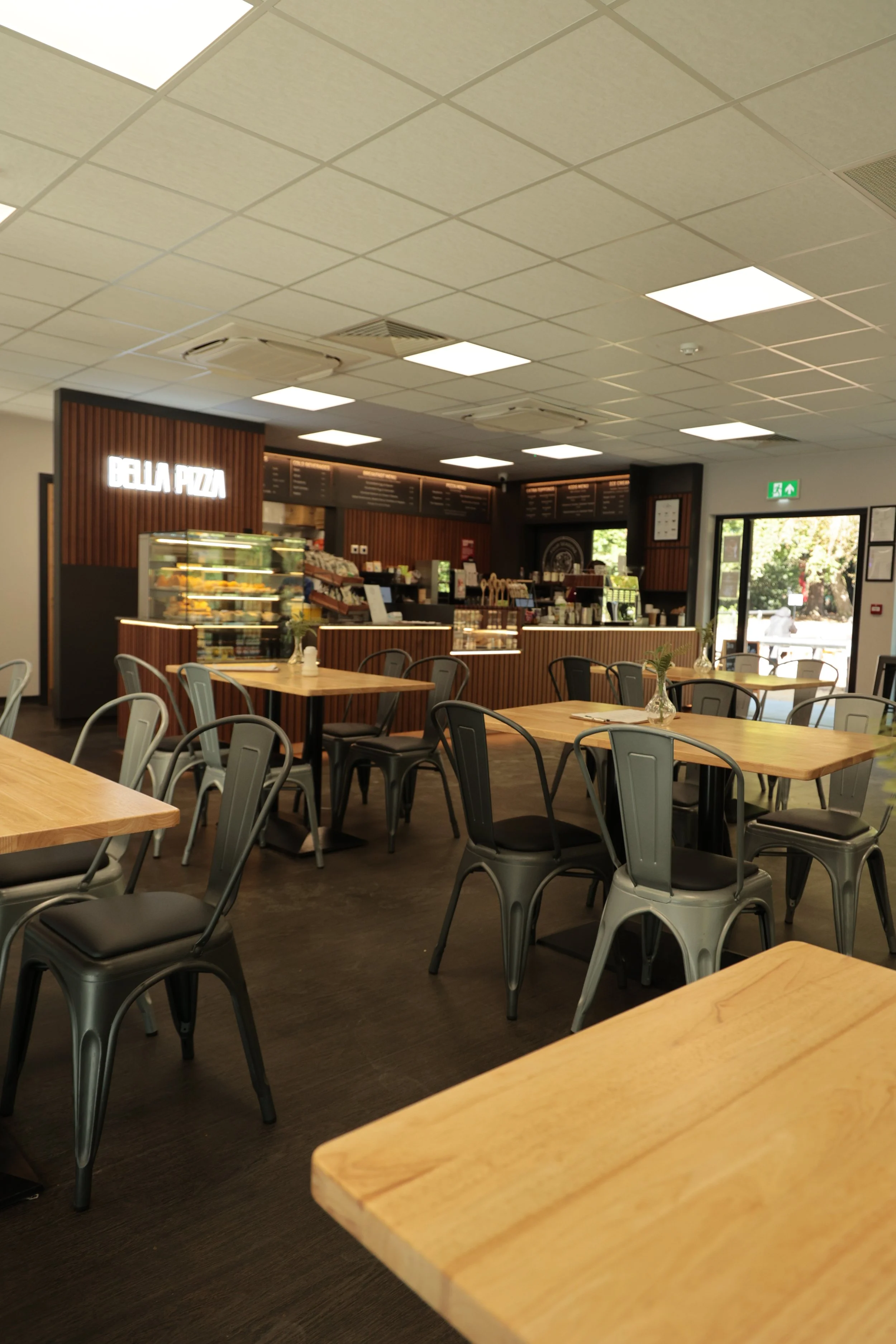 Interior of a modern cafe or restaurant with wooden tables, black and grey chairs, a pizza counter in the background, and large windows letting in natural light.