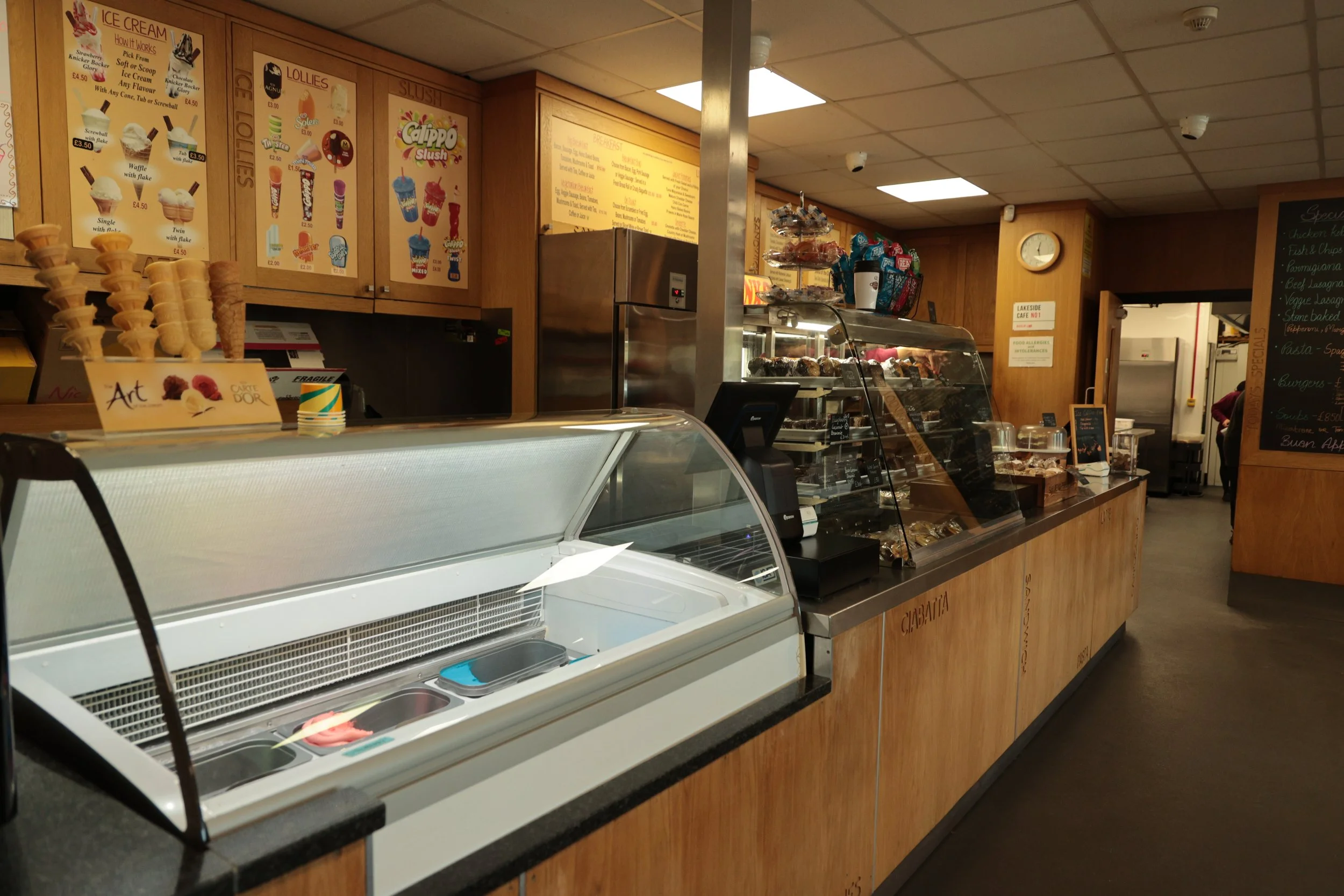 Ice cream counter with various flavors and toppings, menu boards with ice cream options, and a display case with baked goods in a gelato shop.