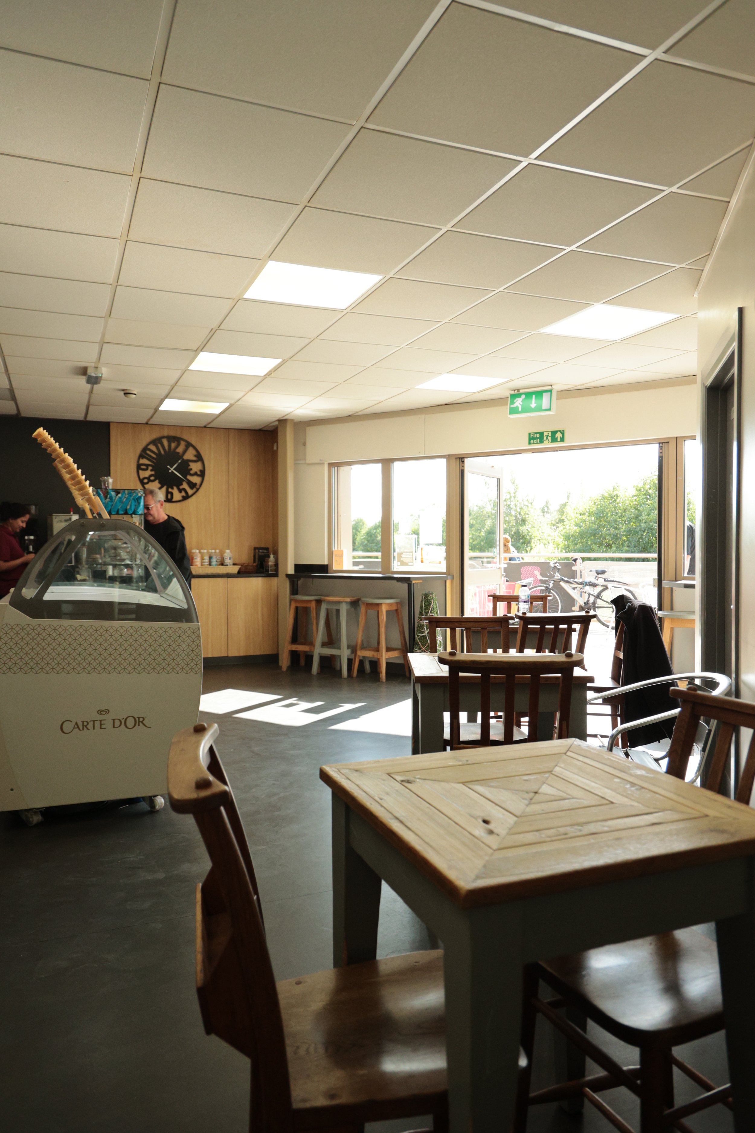 Interior of a cozy café with wooden furniture, an ice cream display, large windows, and natural light.