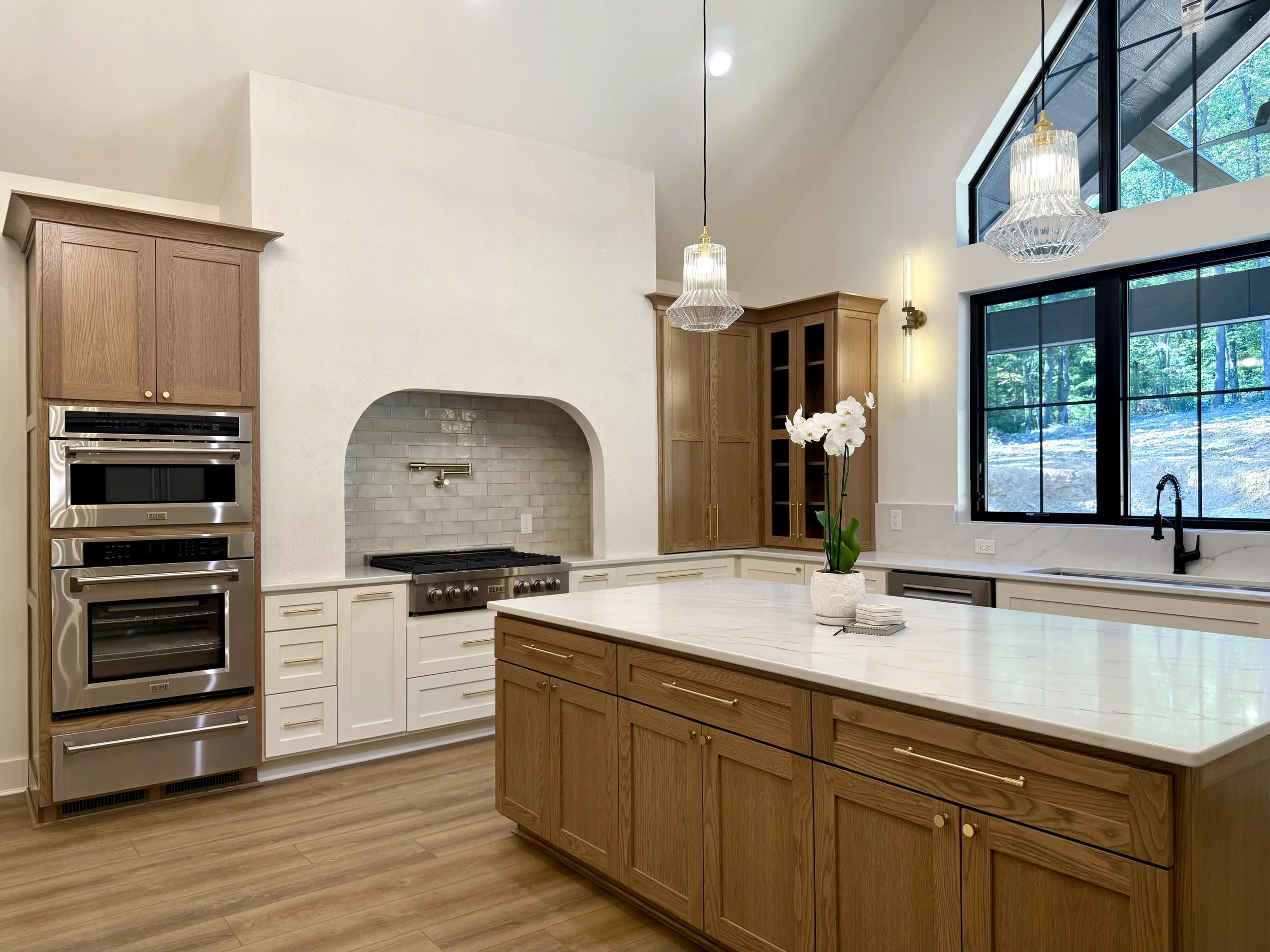 Modern kitchen with wooden cabinetry, quartz island, large window with black framing, pendant lights, and stainless steel appliances.