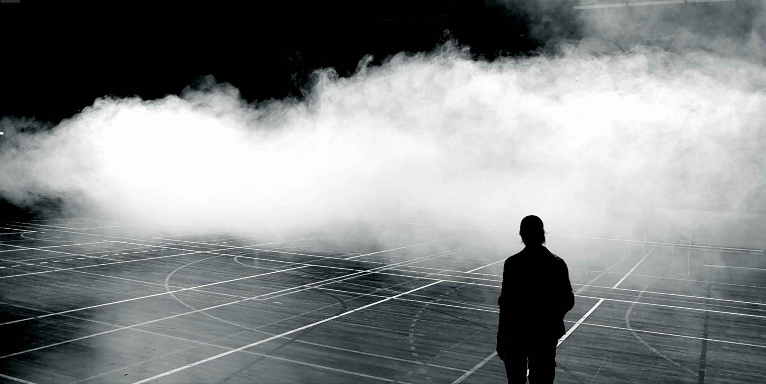 Silhouette of a person standing in a foggy, empty parking lot with fog emitting from a smoke machine or similar source.