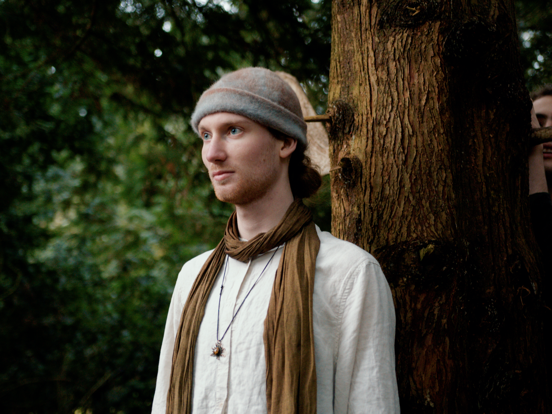 Young man with a beige and brown scarf, wearing a beige shirt and a gray hat, standing outdoors next to a tree in a forested area.