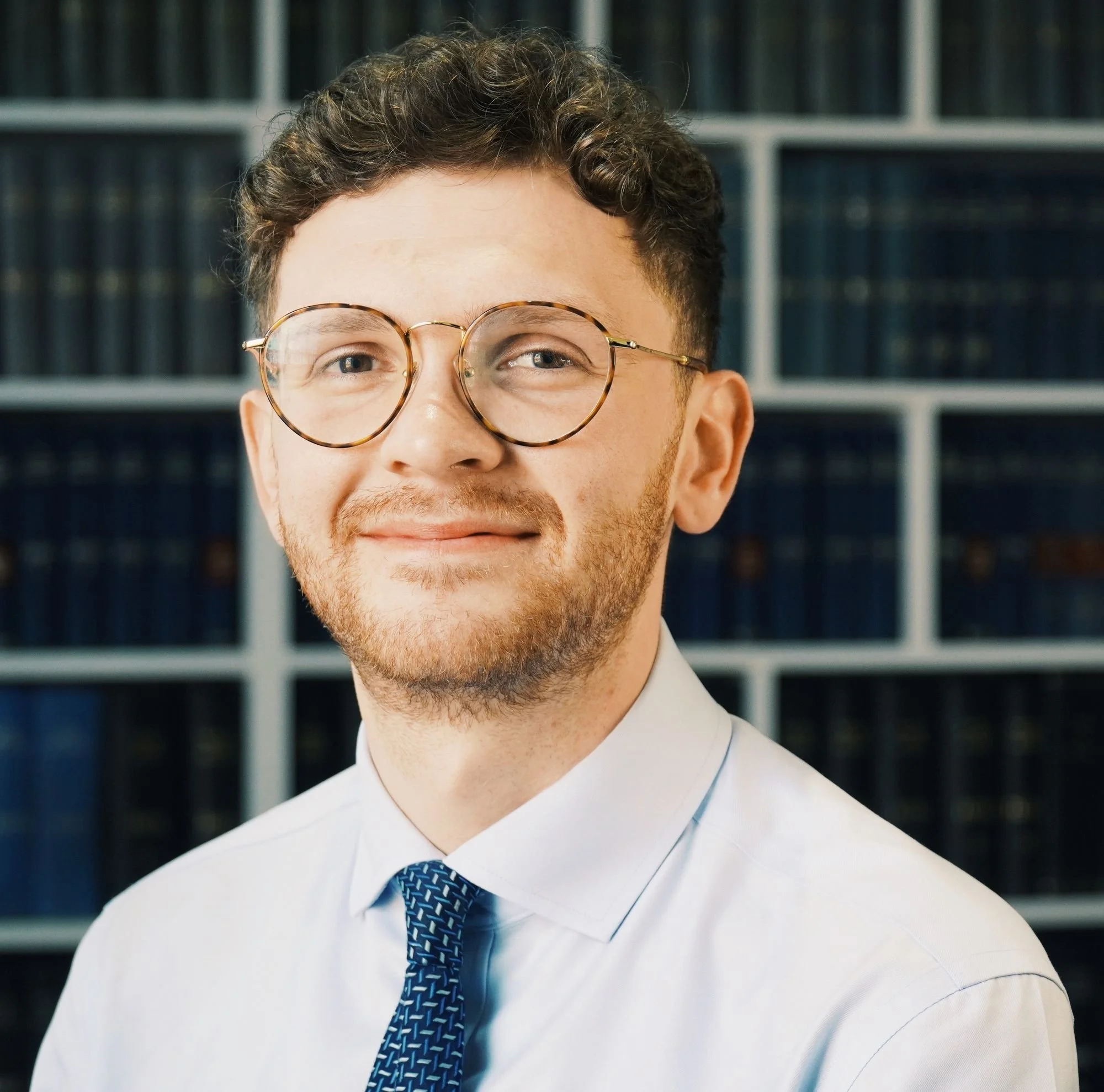 Portrait of a young man with glasses, smiling, in a professional setting with bookshelves in the background.