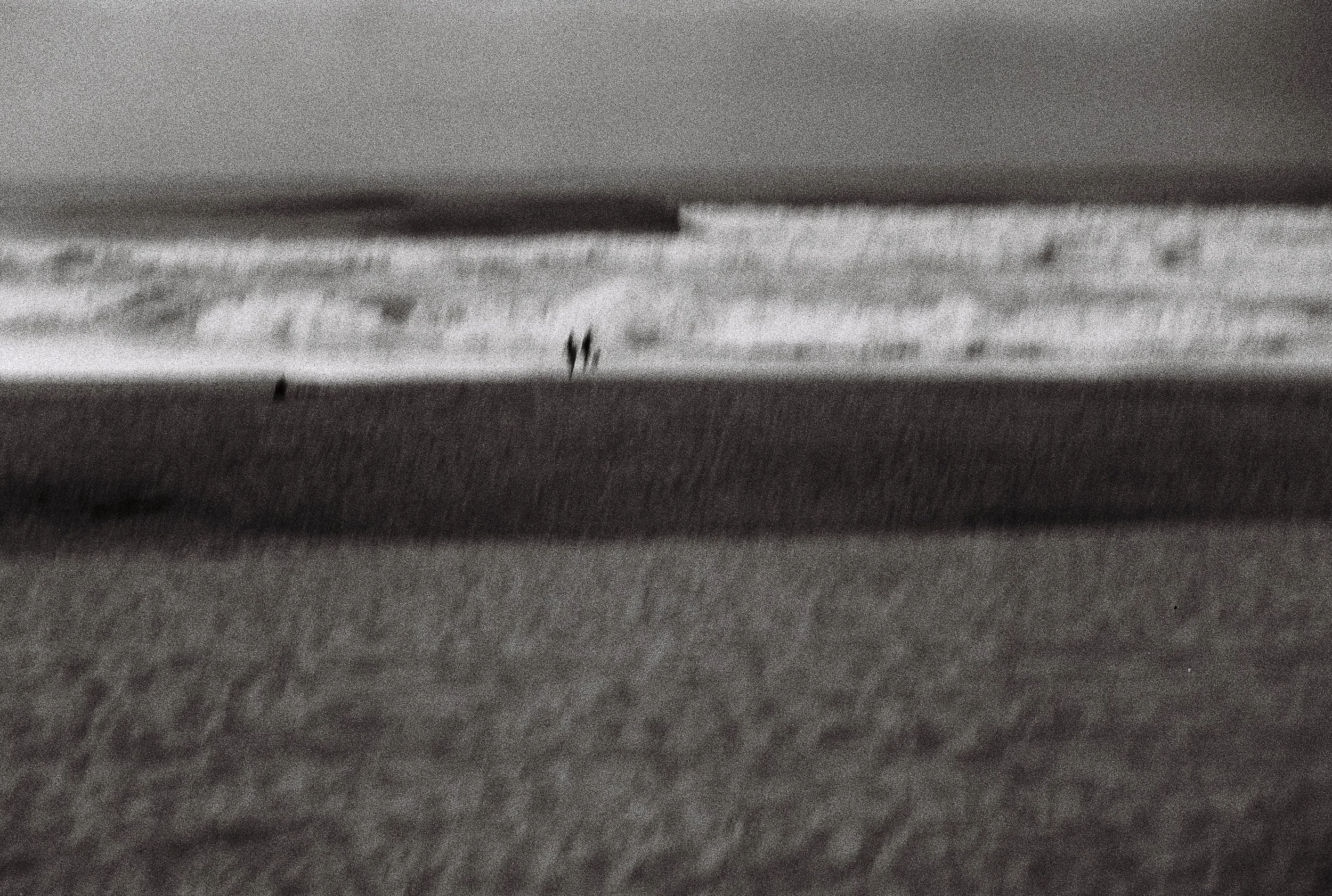 A black and white analog photo of a beach, a surf spot in Hossegor, France, with people standing near the shoreline, waves in the background, and sandy beach in the foreground.