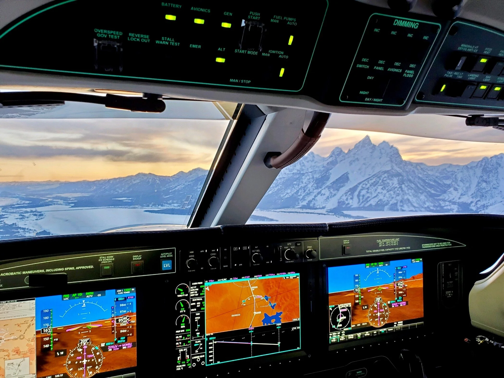 Cockpit of an airplane flying over snow-capped mountains during sunset, with multiple digital screens and controls visible.