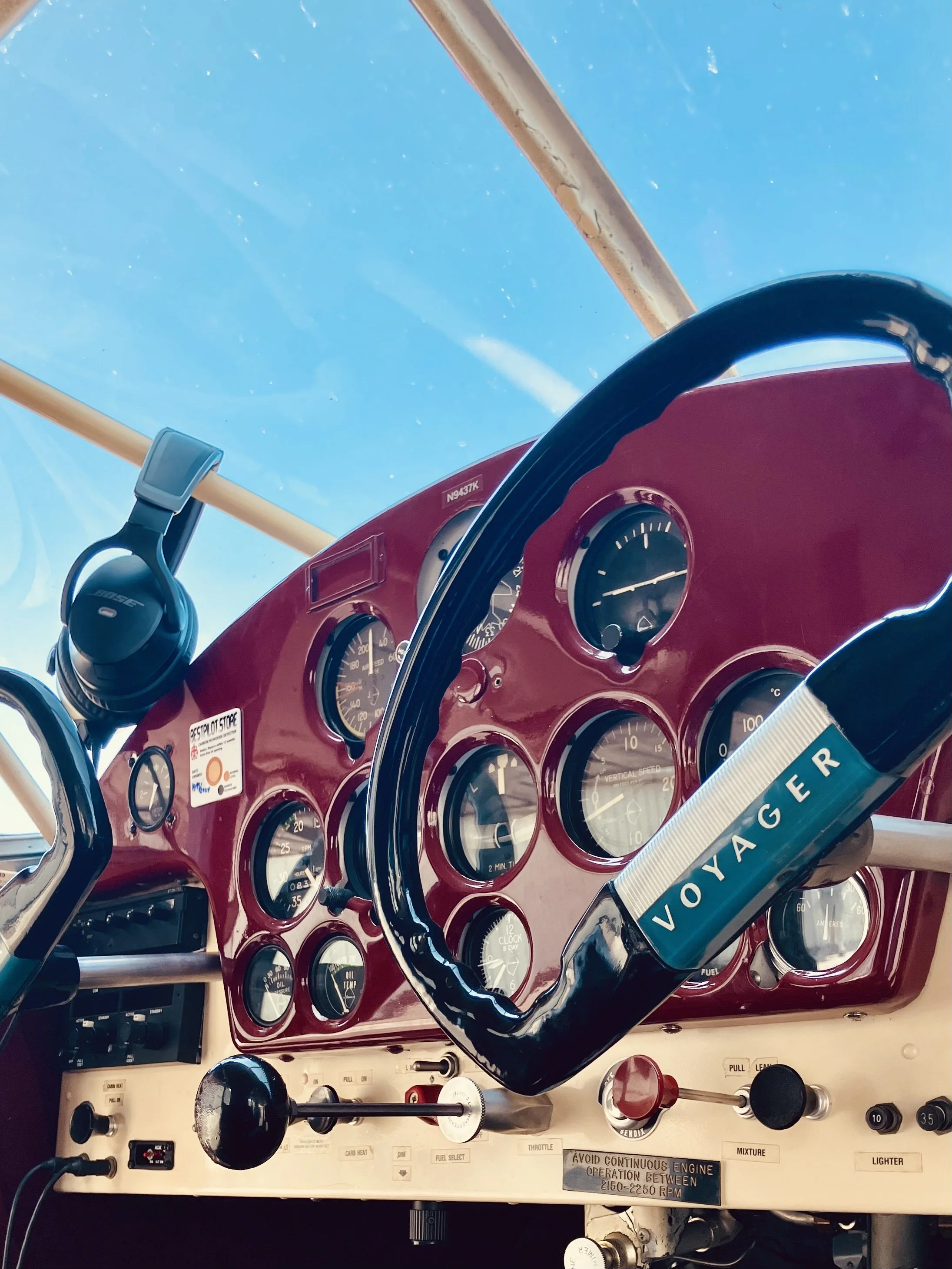 Close-up of an aircraft cockpit's red dashboard with various gauges, controls, and a black Voyager-branded steering wheel. A Bose headset hangs on the side, and a view of the sky is visible through the windshield.