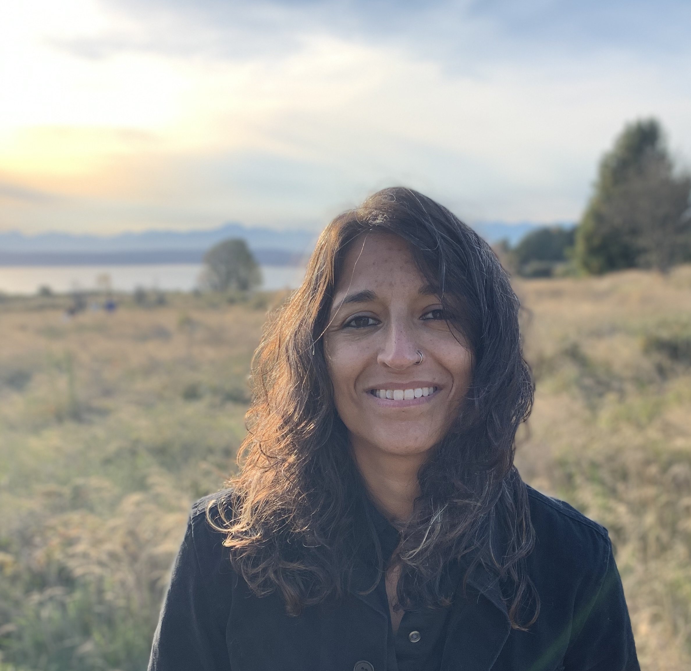 A woman with dark, wavy hair, smiling outdoors at sunset, wearing a black jacket, with a scenic background of water, trees, and mountains.