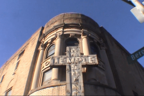 Close-up of an old building with a corner tower, featuring a prominent vertical sign labeled "Temple" and an ornate decorative crest below the sign, under a clear blue sky.