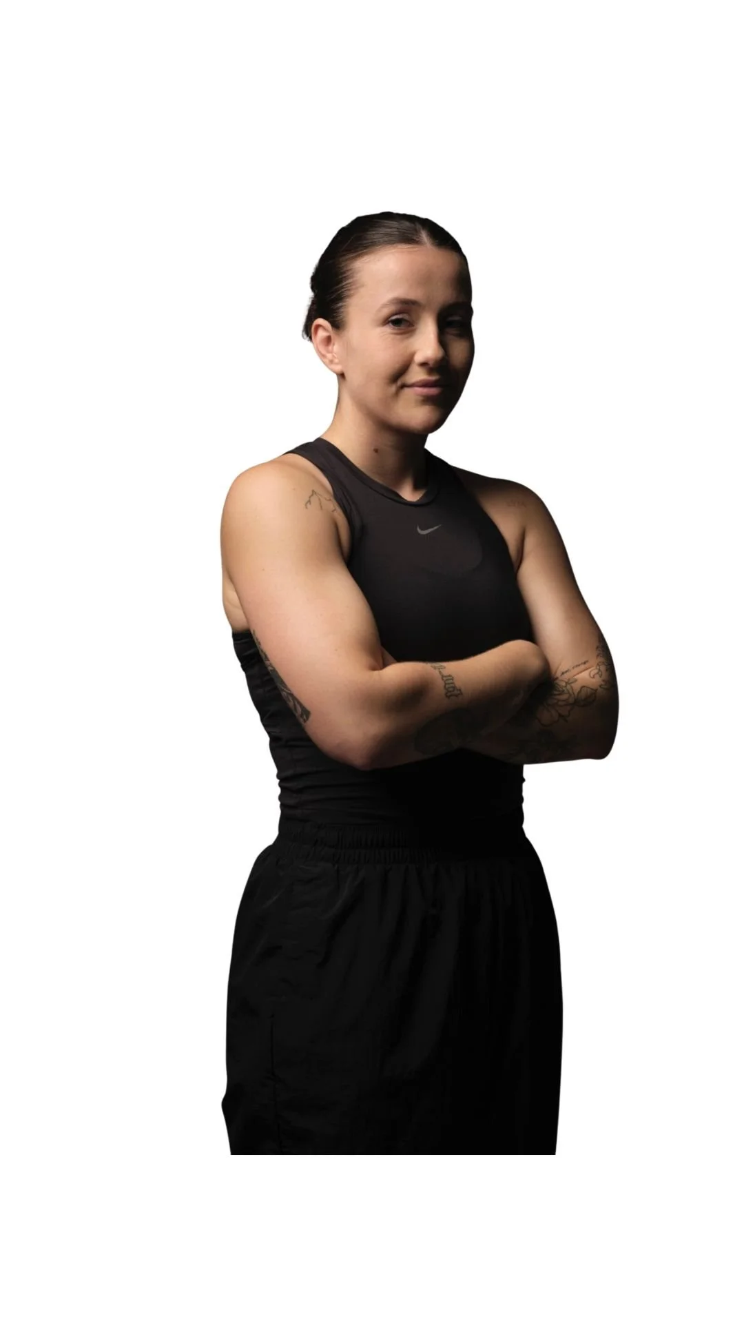 Une femme musclée souriante avec les bras croisés, portant un haut sans manches noir et un pantalon noir, sur fond blanc.