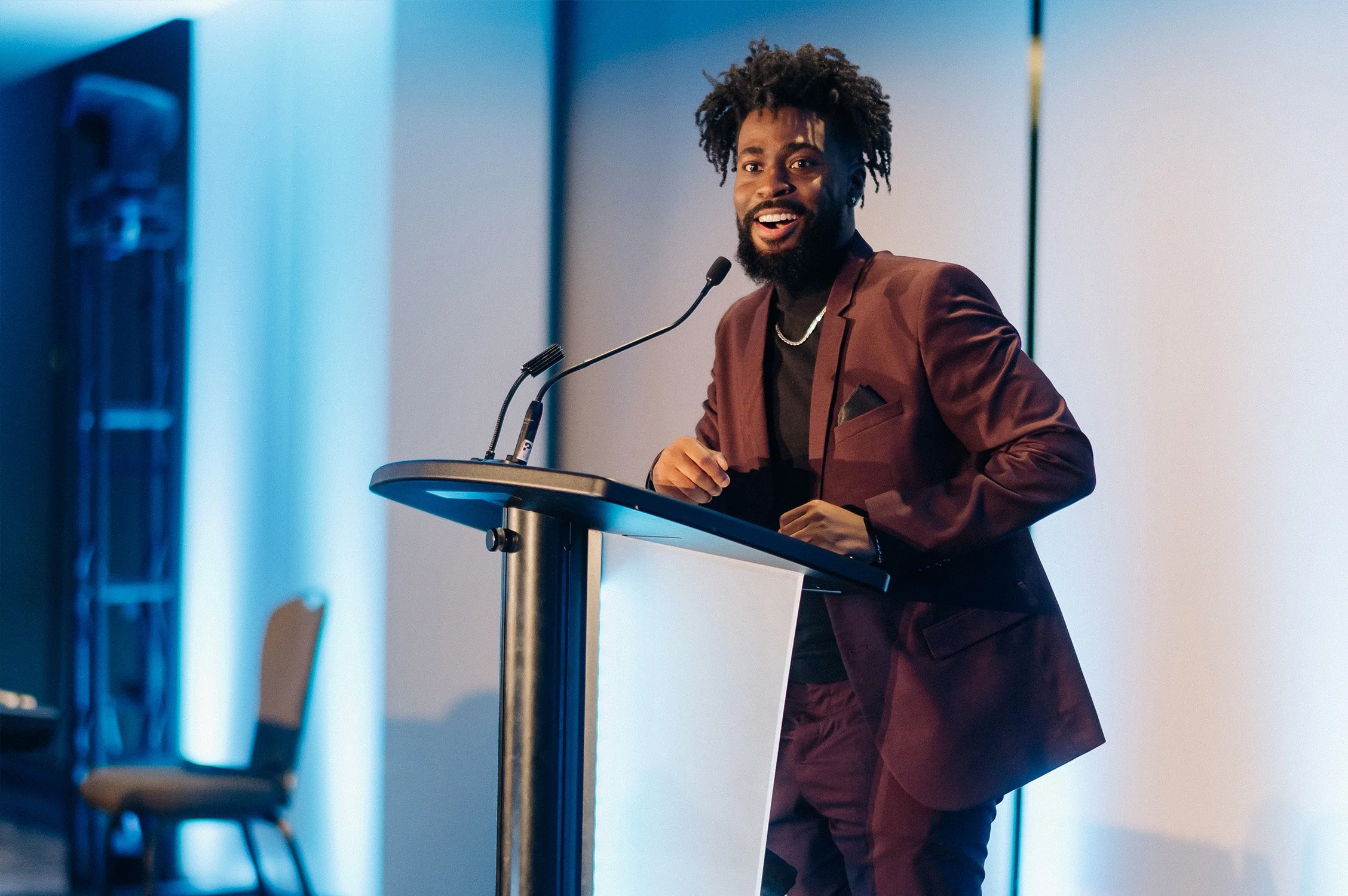 A man in a burgundy suit and black shirt is smiling and speaking at a podium with a microphone.