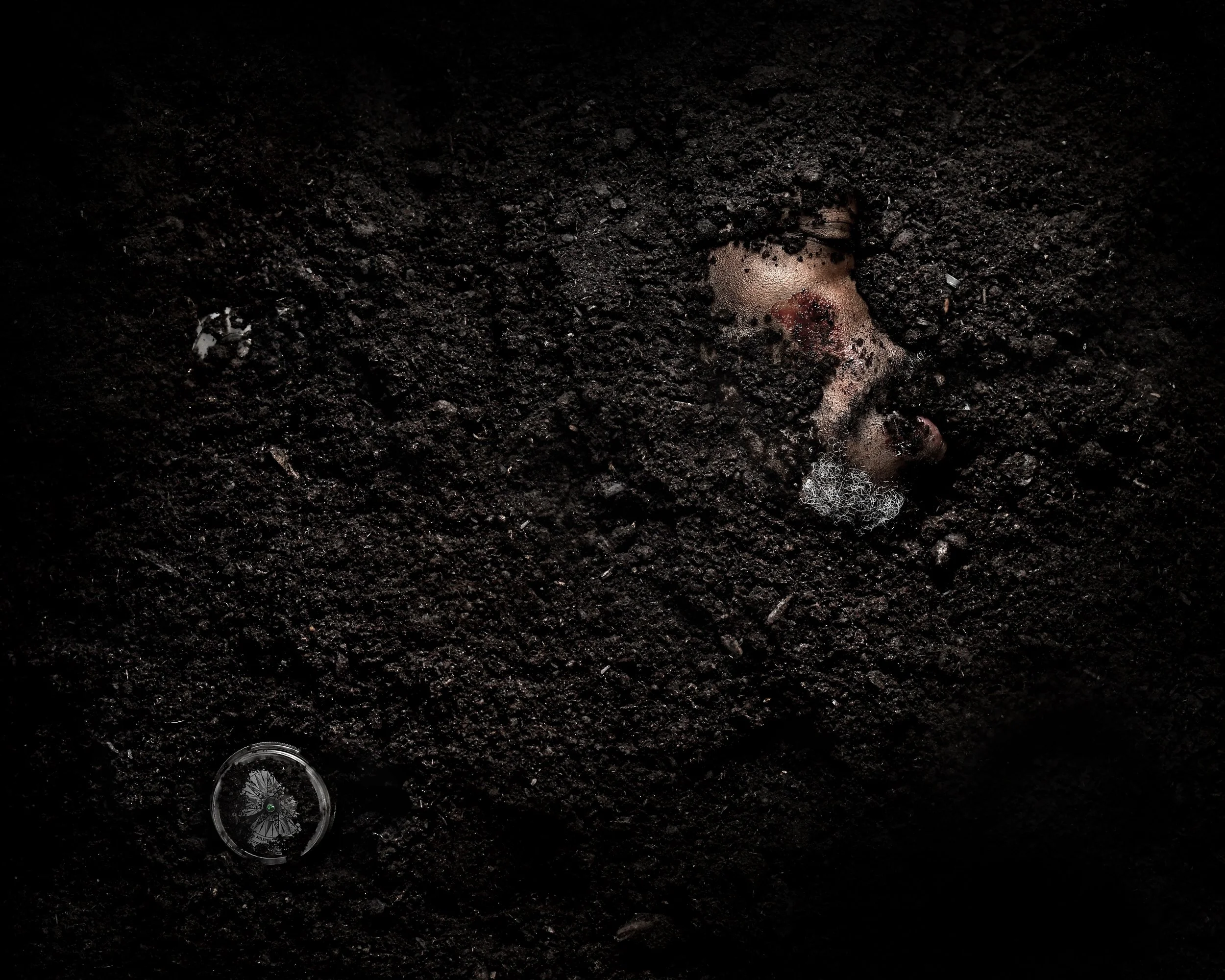 A human face partially buried in dark soil, with the eyes and nose visible, and a small round container on the soil nearby.
